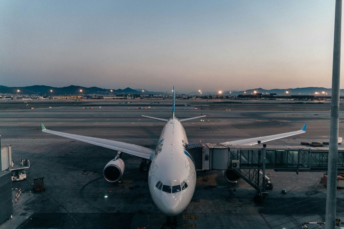 A plane at Barcelona Airport connected to a jet bridge with a runway and mountains in the background