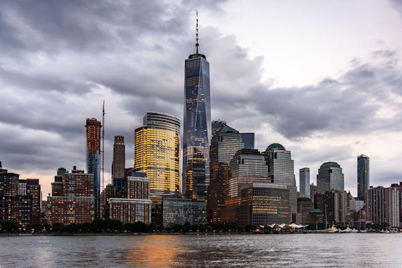 A view of New York City’s skyline with the One World Trade Center, reflecting lights under a cloudy sky