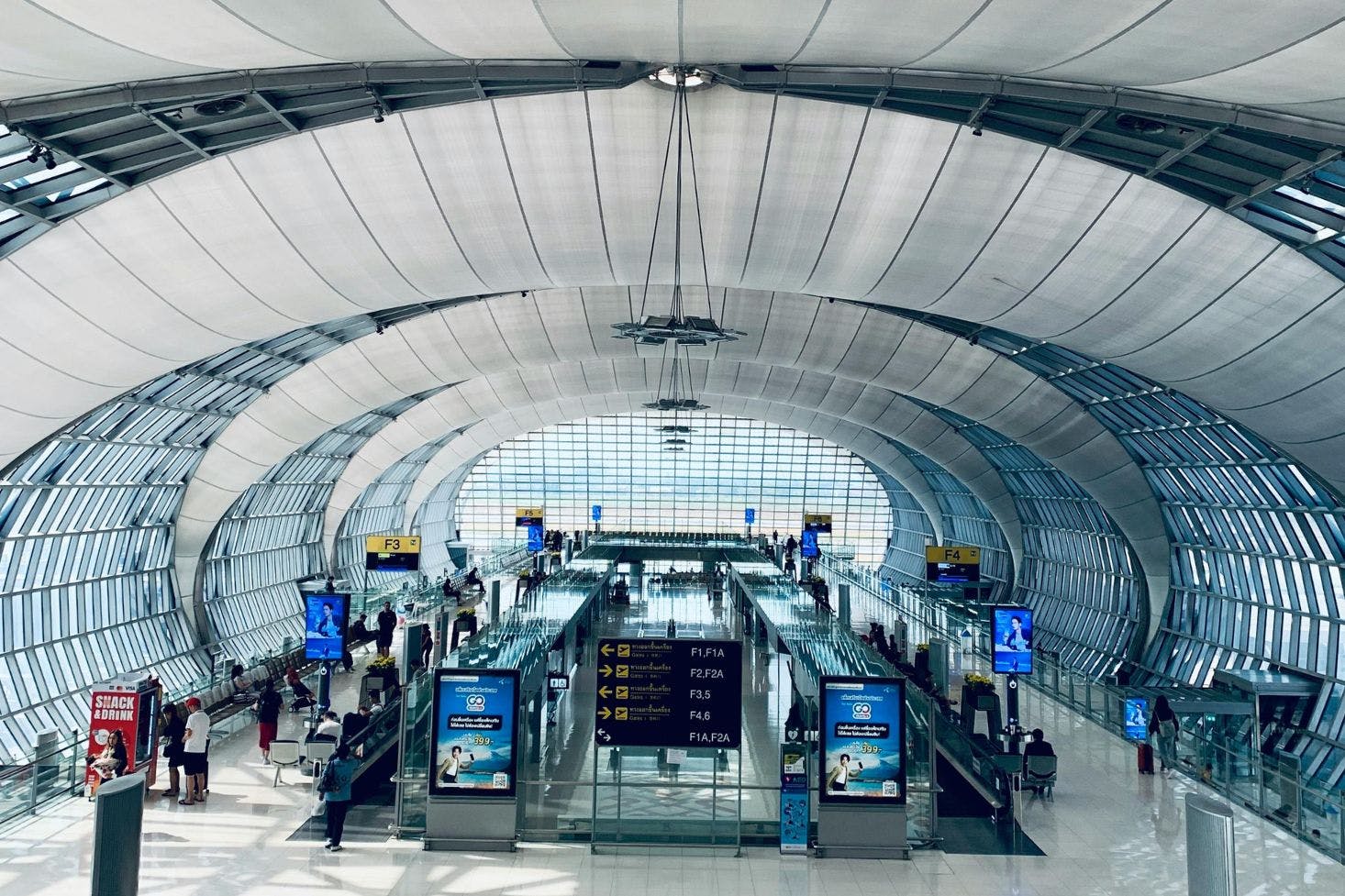 Oval-shaped glass hall at Suvarnabhumi Airport, Bangkok, with gates, signs, seating area, and passengers