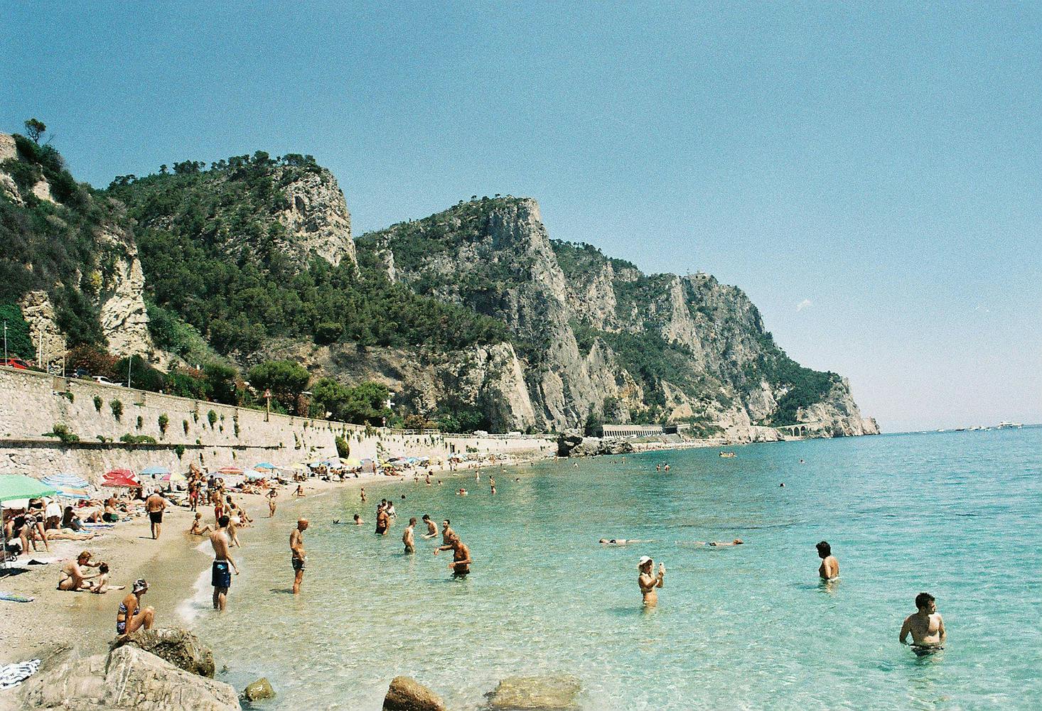 Bagnanti al mare a Savona, con acque cristalline e sabbia dorata in primo piano, e costa rocciosa e cielo azzurro sullo sfondo 