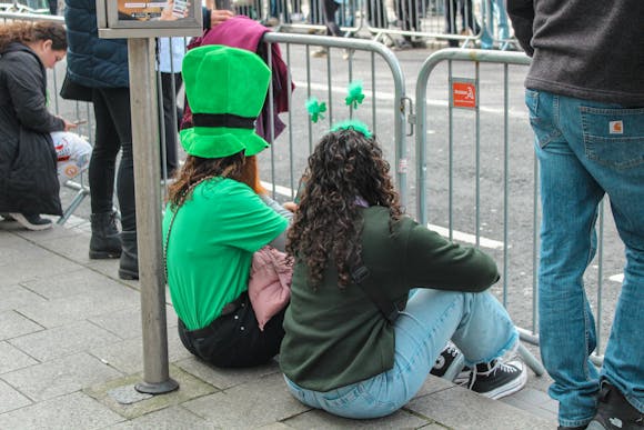 Two parade attendees dressed in festive green attire, including a leprechaun hat and shamrock headband, waiting for the St. Patrick's Day Parade