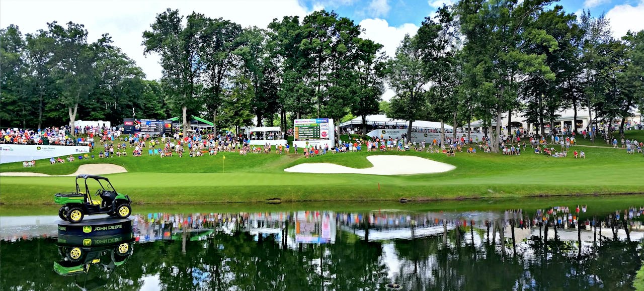 Spectators lined up along a green golf course beside a body of water, enjoying the action at the Farmers Insurance Open