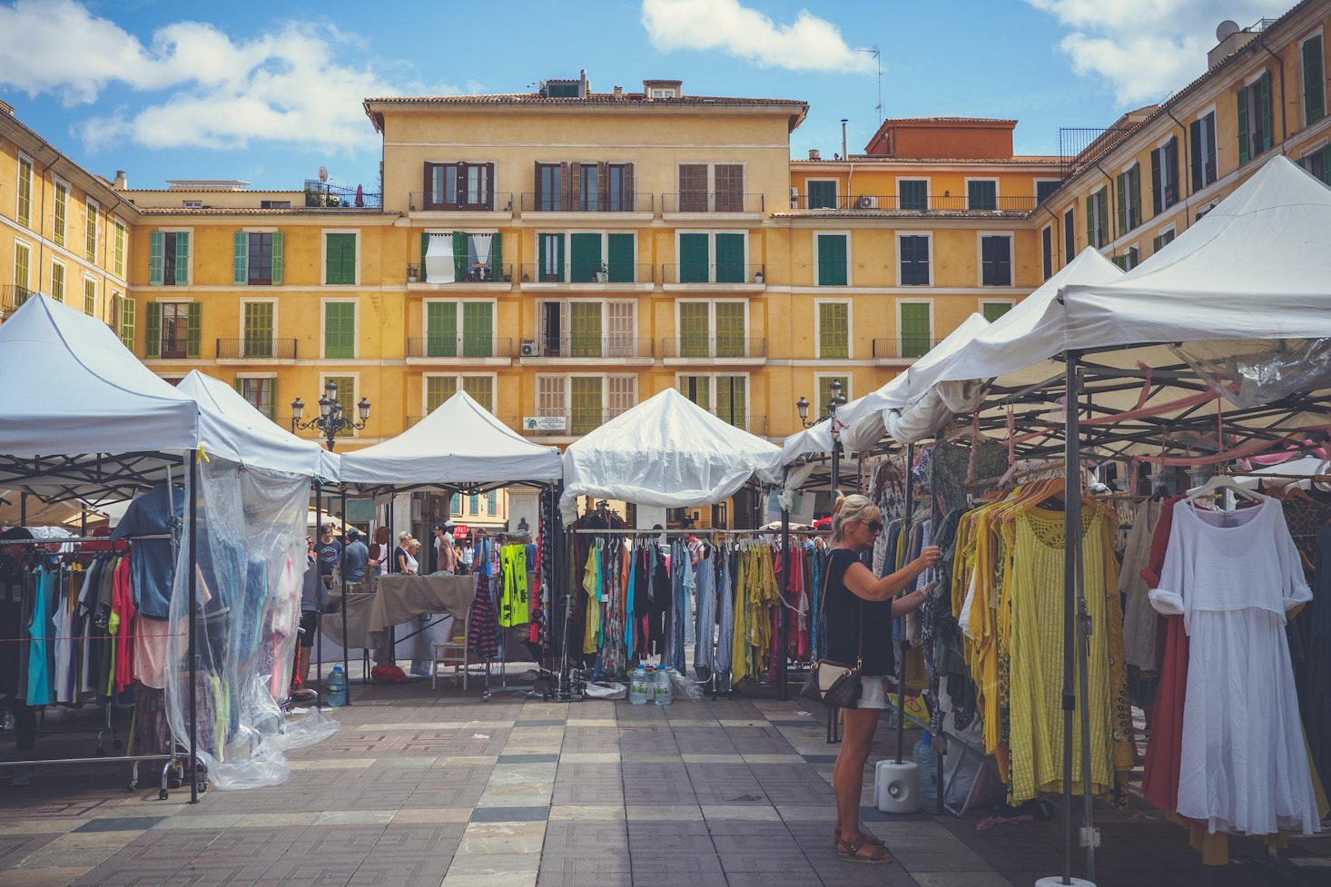 Blonde tourist browsing colorful clothing stands at Plaça Major, a market square in Palma de Mallorca