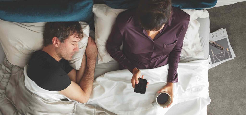 Man and woman laying in bed, woman is holding her phone and cup of coffee