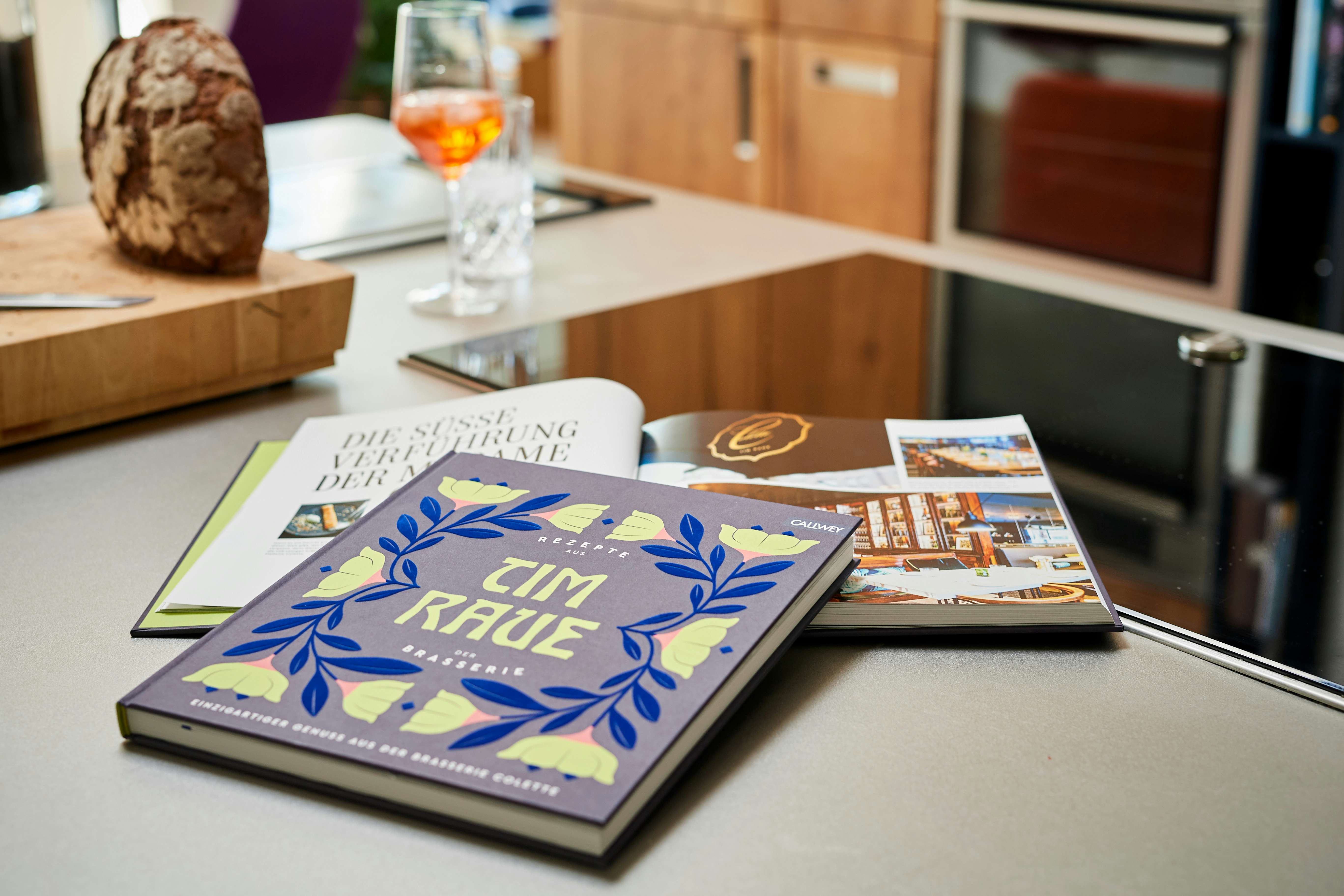 Das Kochbuch »Tim Raue – Rezepte aus der Brasserie« auf der Küchenarbeitsfläche. Ein Glas Apperol und ein Laib Brot im Hintergrund.