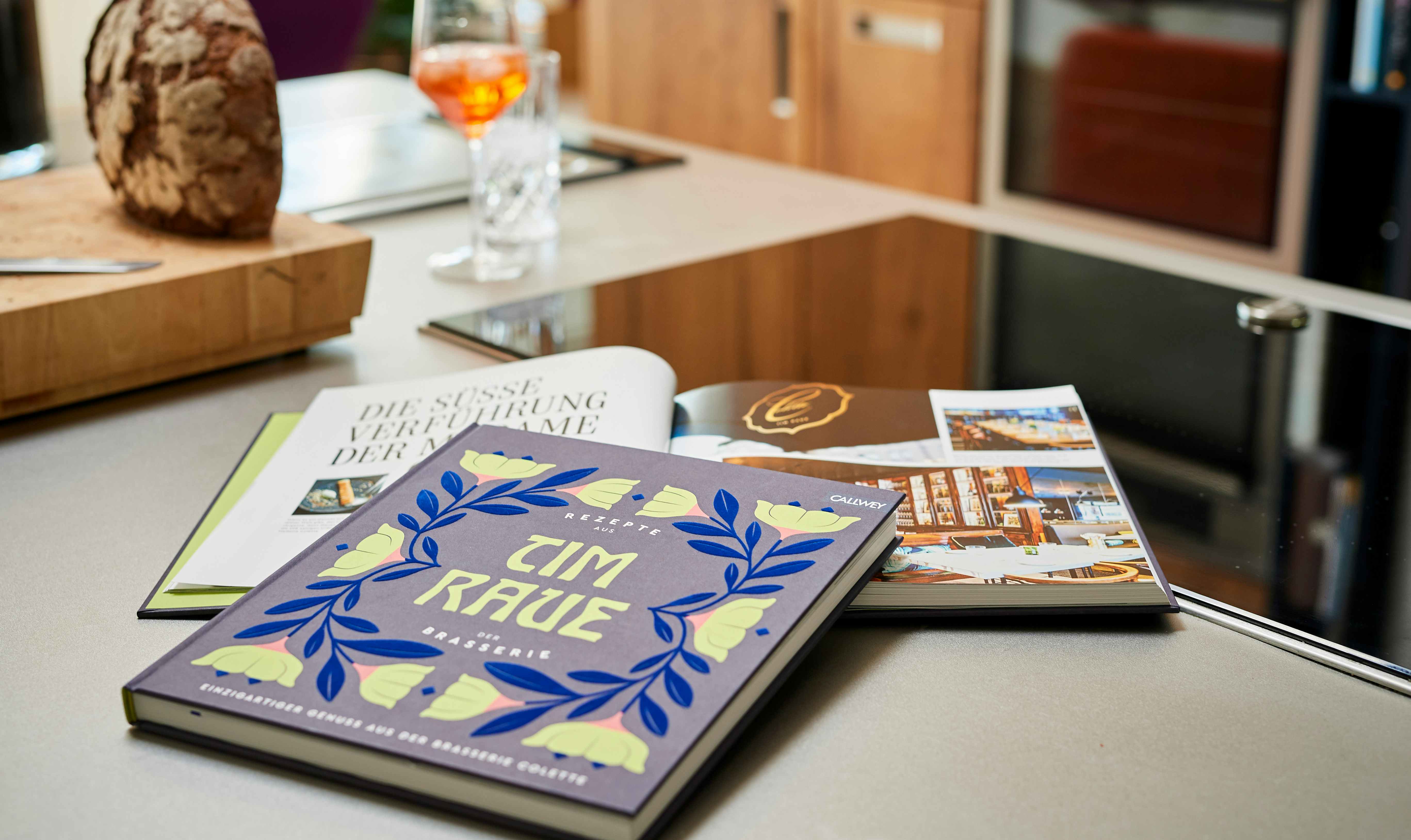 Das Kochbuch »Tim Raue – Rezepte aus der Brasserie« auf der Küchenarbeitsfläche. Ein Glas Apperol und ein Laib Brot im Hintergrund.