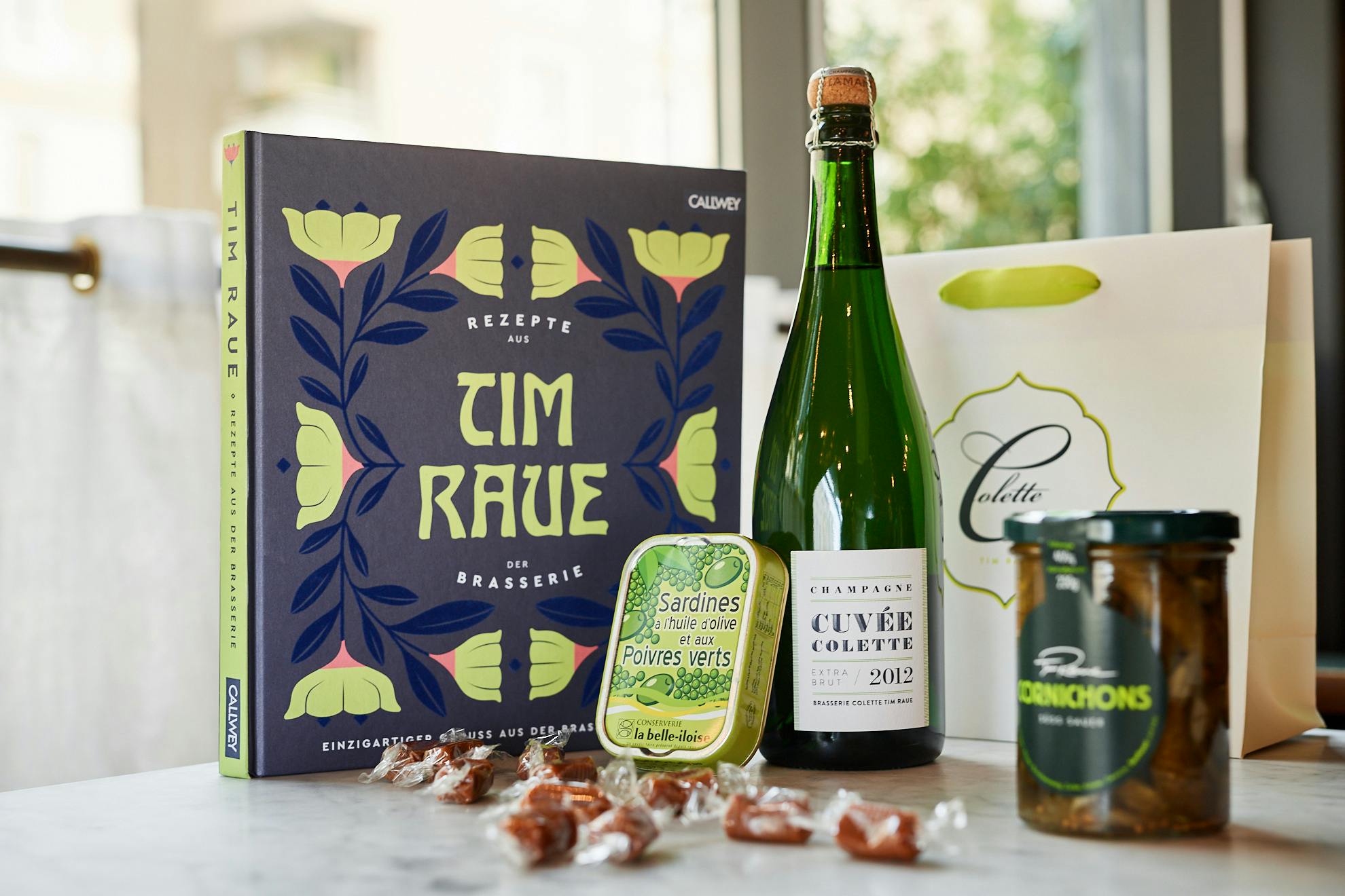 Produkte aus der Brasserie Colette: Kochbuch von Tim Raue, Sardinenbüchse, Champagner-Flasche, Cornichons und Papiertasche