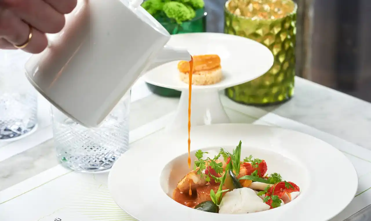 Angerichtete Speise: Bouillabaisse, Kellner gießt die Fischsuppe aus einem weißen Kännchen an
