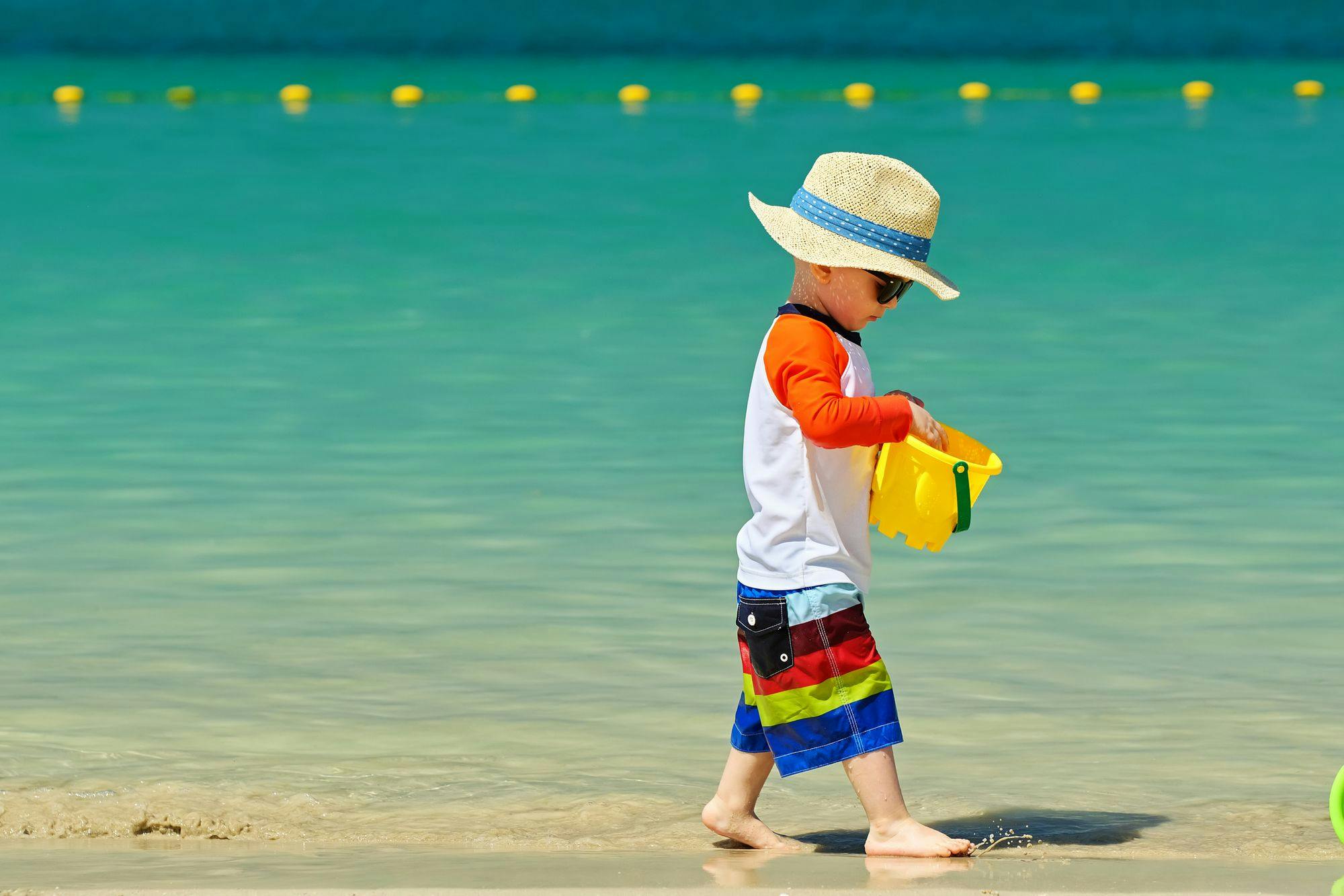 Small child wearing a hat and sunglasses, carries a pail while walking along the shore at a beach.