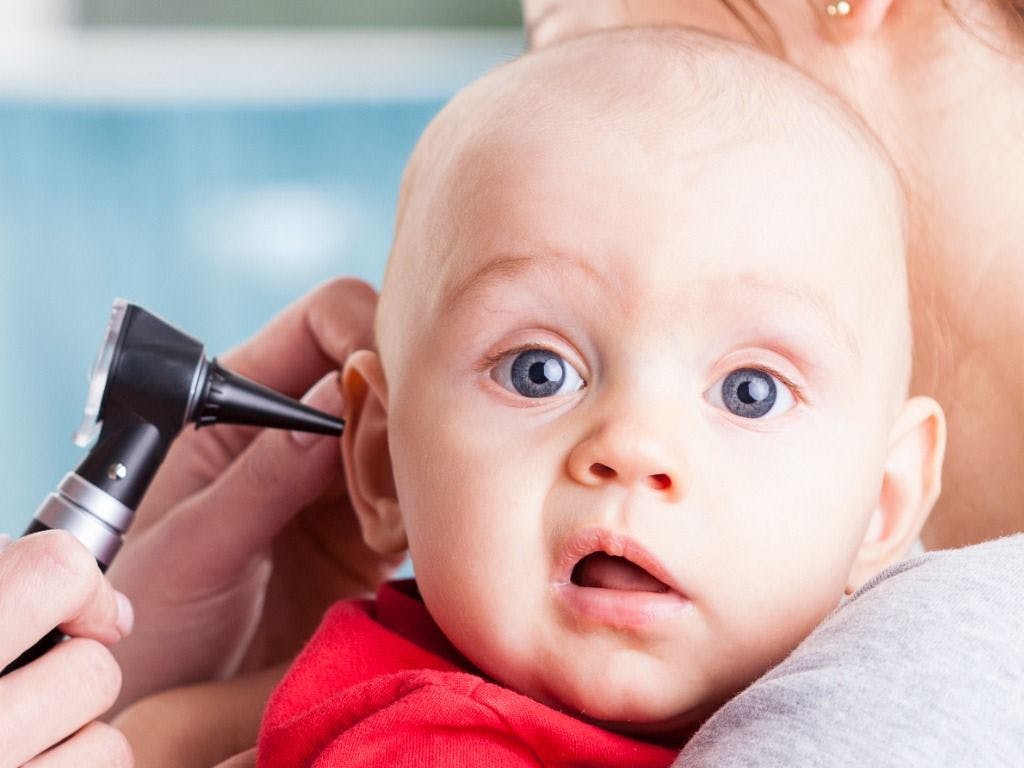 Bald headed, blue eyed baby with an otoscope in the ear. 