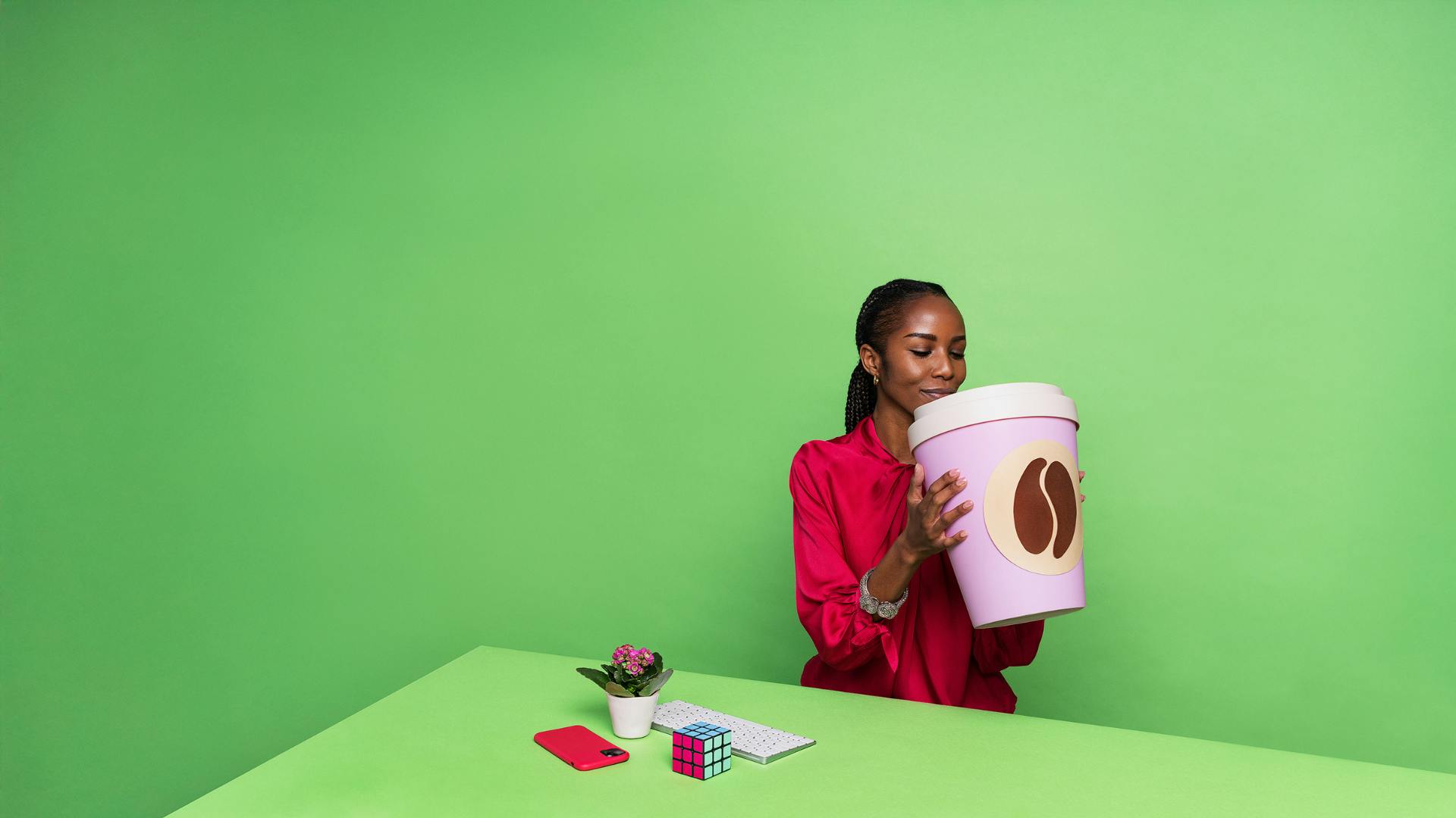 En person i rosa kläder sitter bakom ett grönt skrivbord framför en grön bakgrund. PÅ bordet finns ett tangentbord, en rosa mobiltelefon en rubiks kub, en blomma i en kruka och en jättestor kaffemugg.