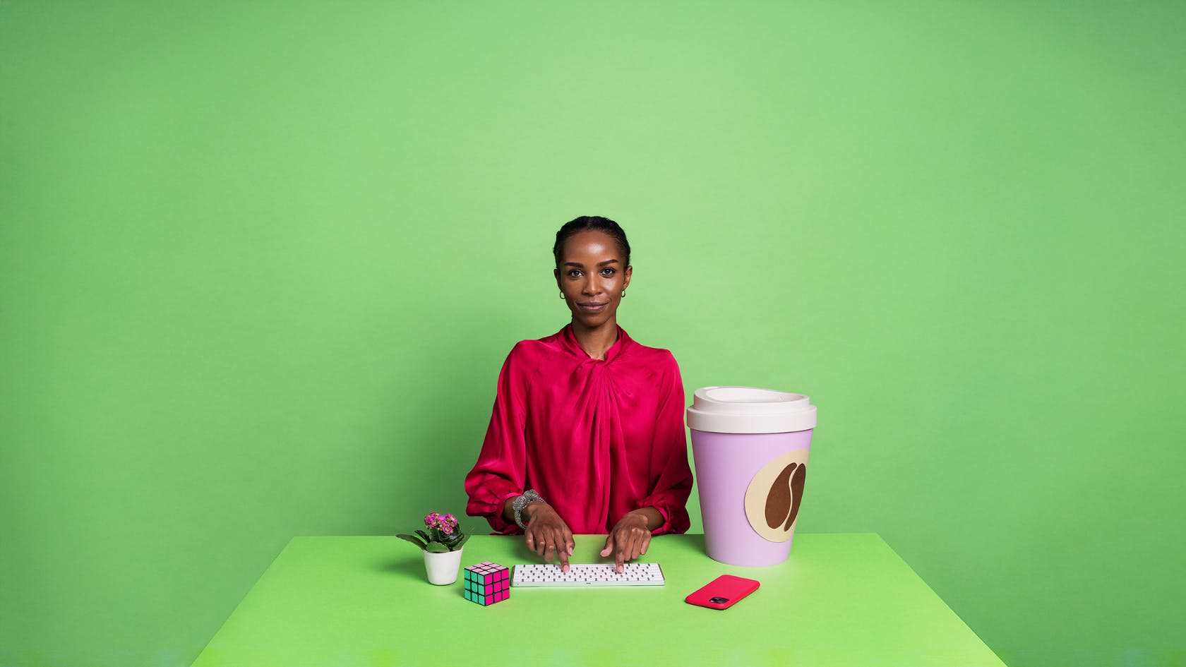 En person i rosa kläder sitter bakom ett grönt skrivbord framför en grön bakgrund. PÅ bordet finns ett tangentbord, en rosa mobiltelefon en rubiks kub, en blomma i en kruka och en jättestor kaffemugg.