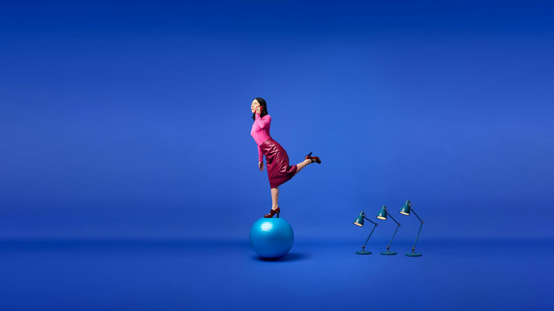 En person i rosa kläder står på ett ben på en stor pilatesboll samtidigt som hen pratar i telefon