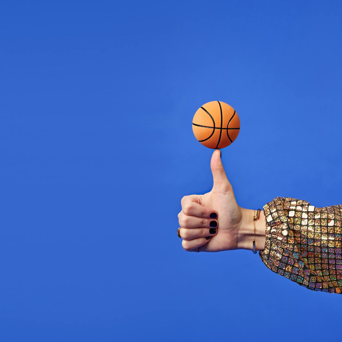 En tumme som balanserar en liten basketboll på tummen. Guldglittrig tröja. Blå bakgrund.
