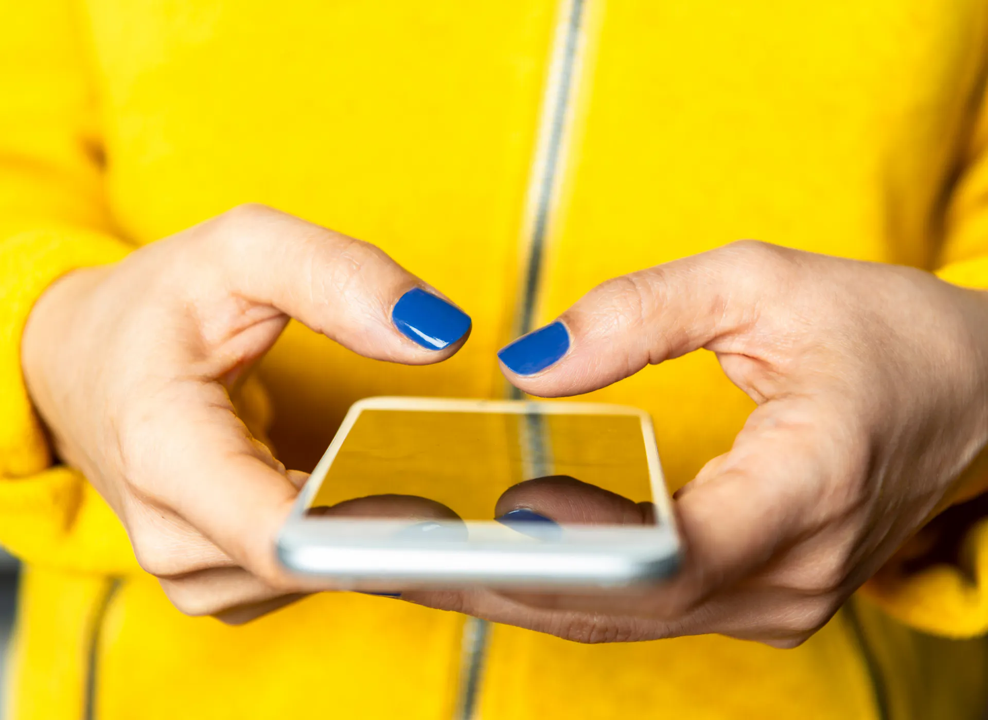 Inzoomat på en person som håller i en mobiltelefon med båda händerna. Gul tröja och blått nagellack.