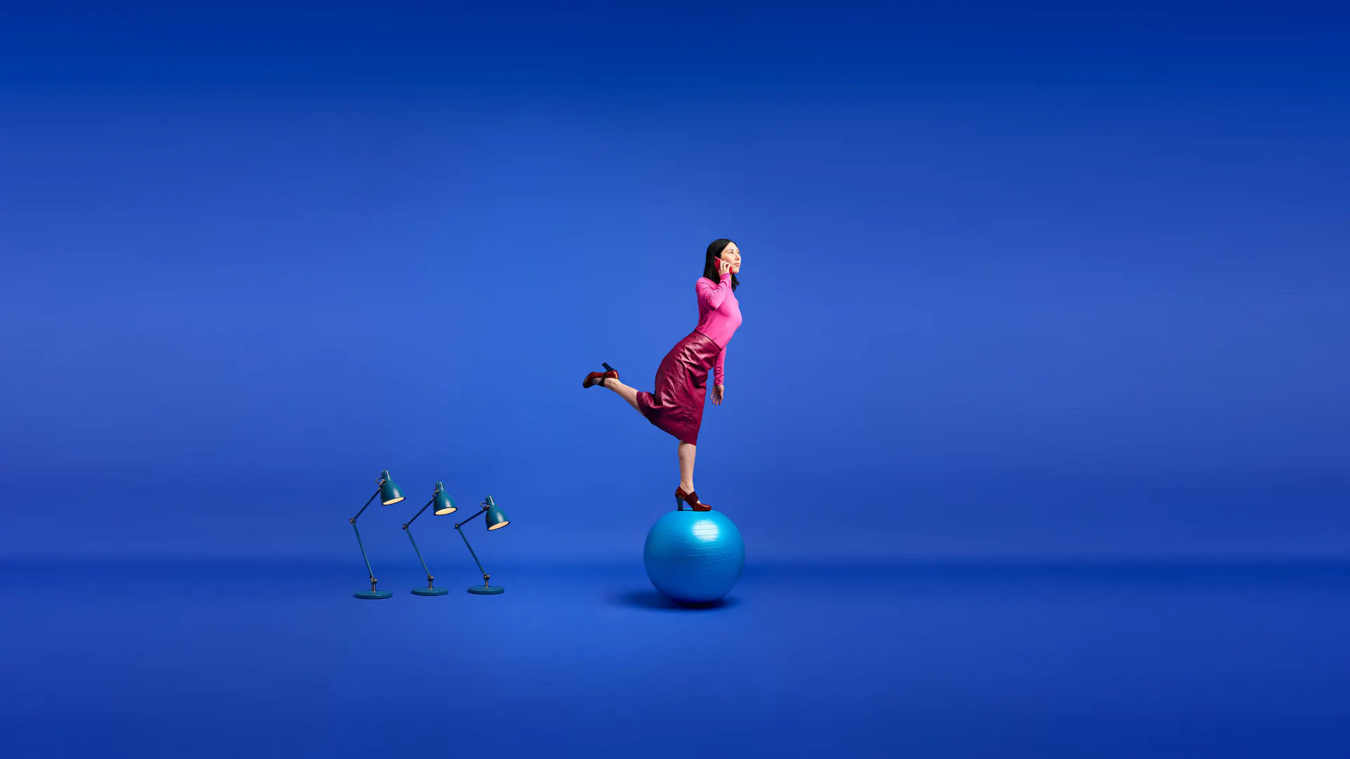 Person balanserar på en blå pilatesboll mot blå bakgrund, med en telefon mot örat och tre skrivbordslampor i bakgrunden.