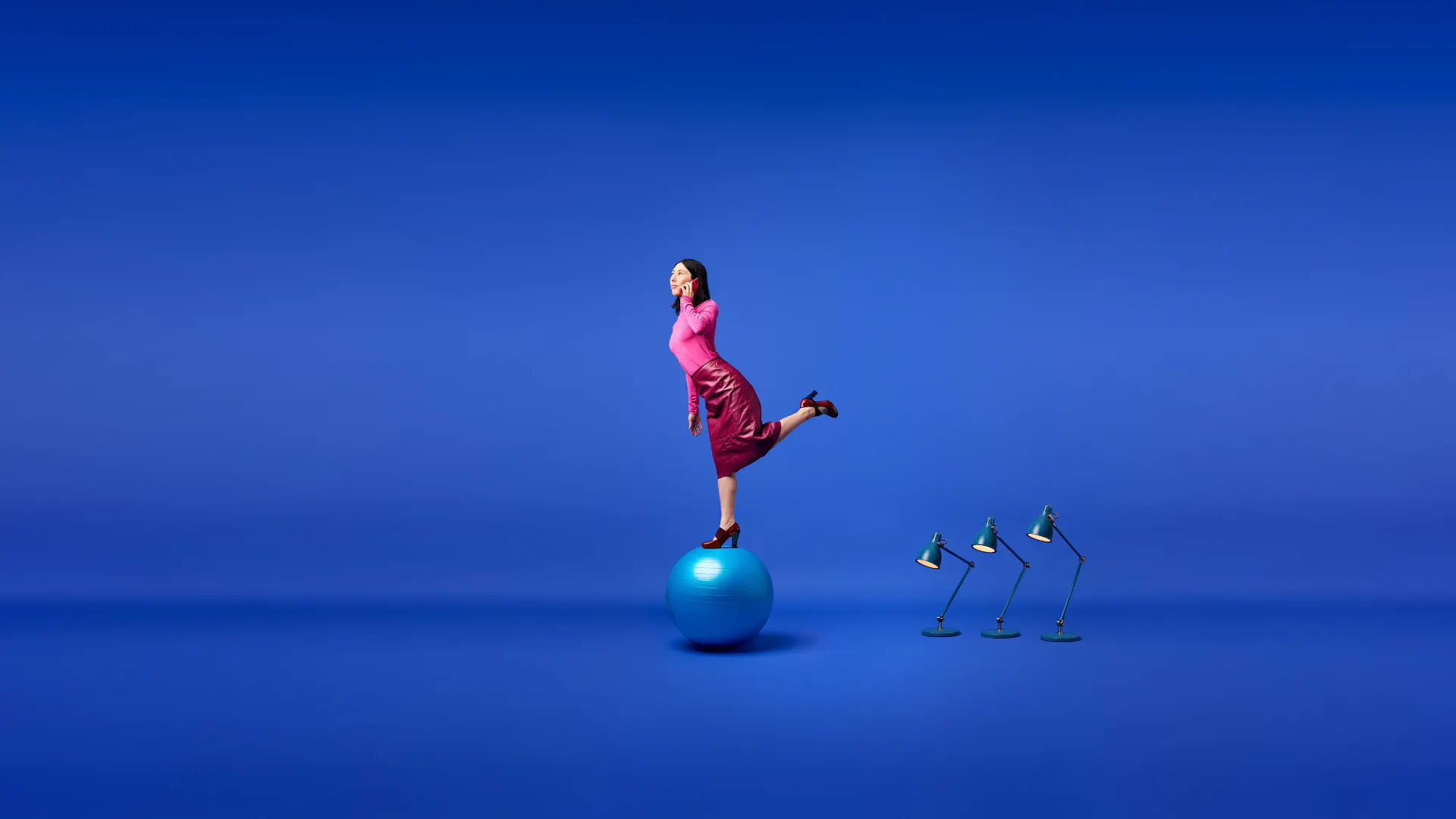 En person balanserar på en stor blå träningsboll mot en helblå bakgrund. Personen bär rosa kläder och står på ett ben med ena handen vid sidan av huvudet. Till höger om bollen står tre skrivbordslampor på rad, lutade i samma riktning. Bilden har ett minimalistiskt uttryck med starka kontraster mellan den blå bakgrunden och de rosa tonerna i kläderna.