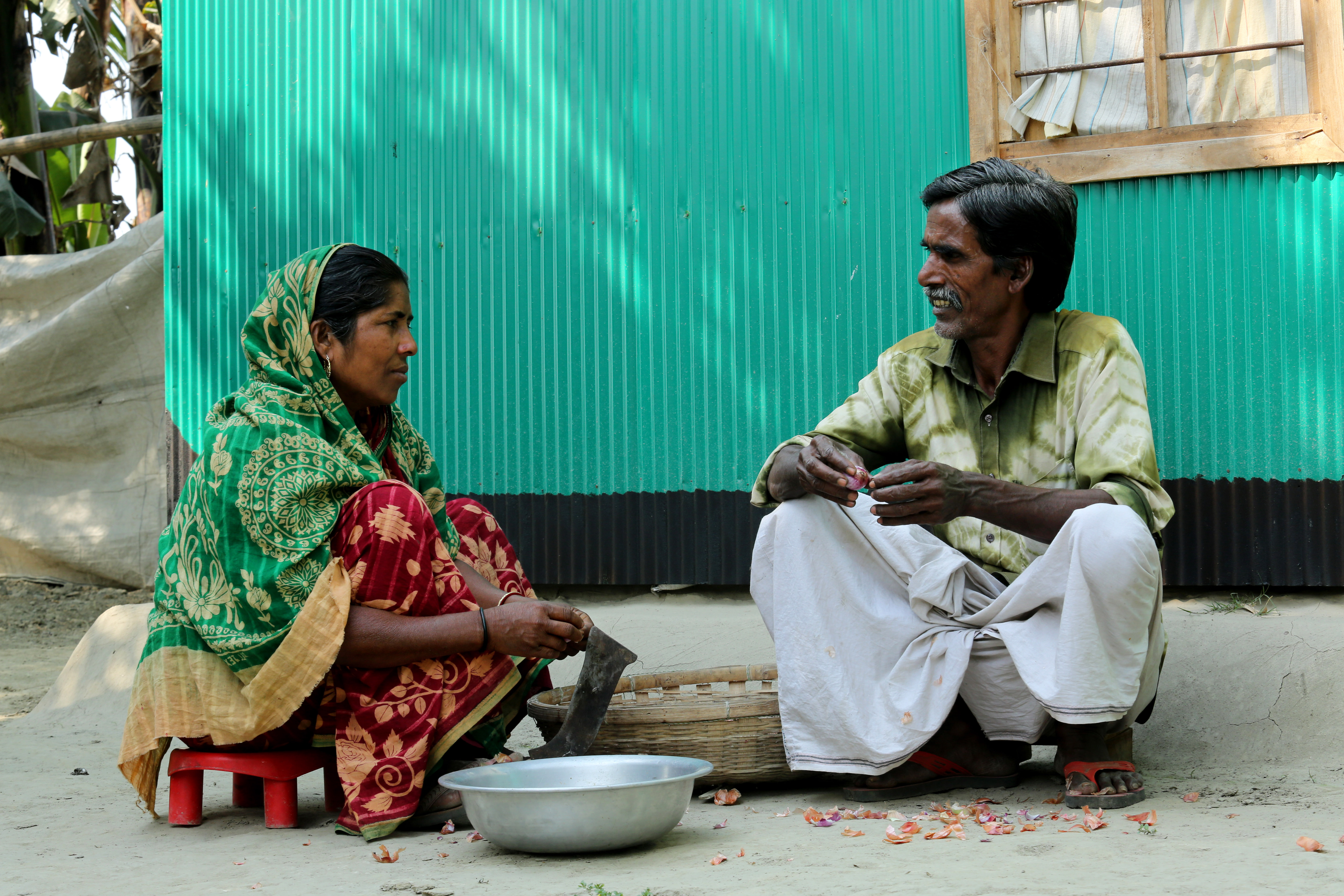 Picture of Fulchan (right) wearing white trousers and green shirt, and Joynab (left) wearing a green sari.  They are pictured sat outside their house in Bangladesh.