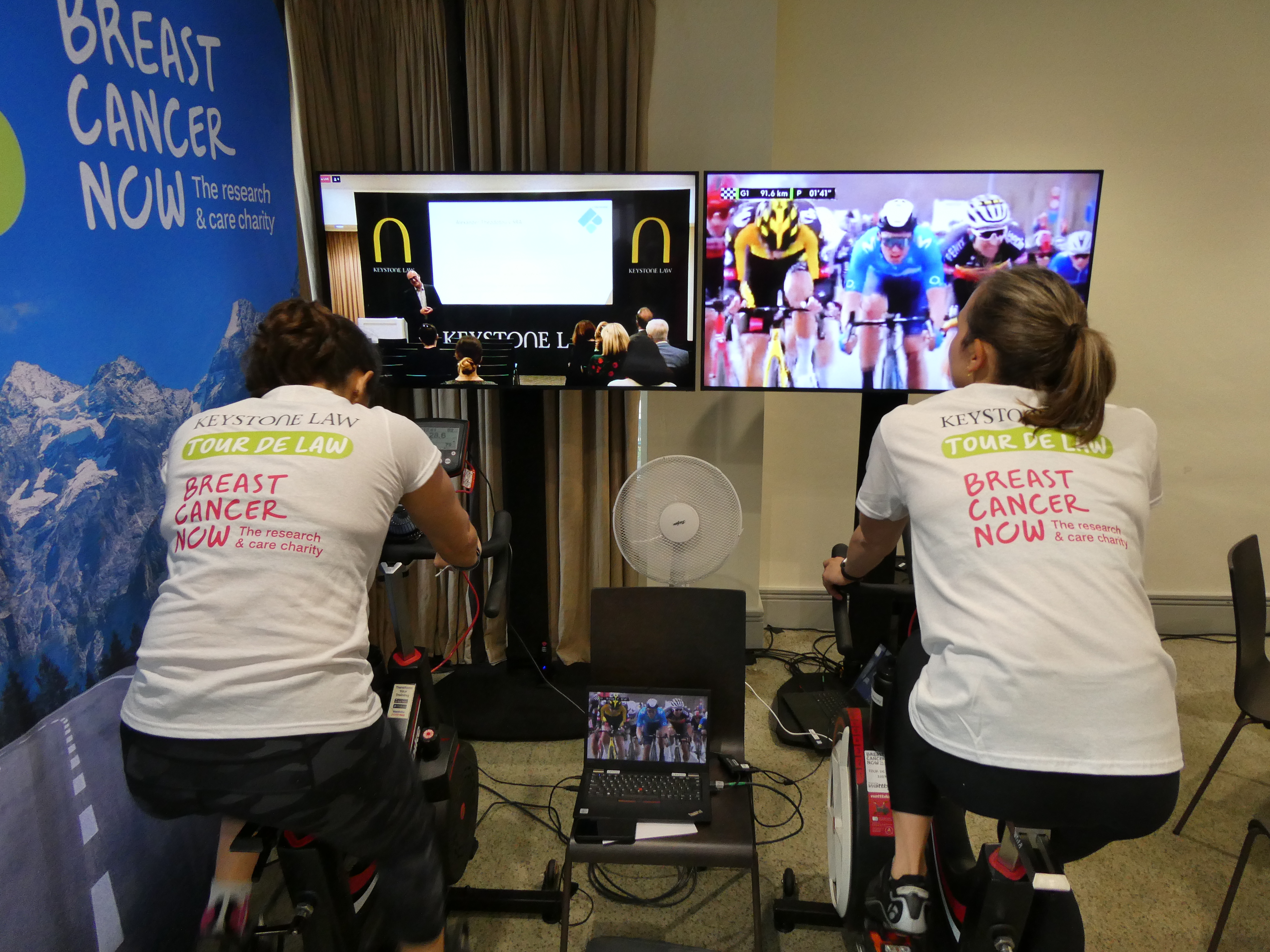 Two cyclists on static bikes facing away from the camera towards TV screens