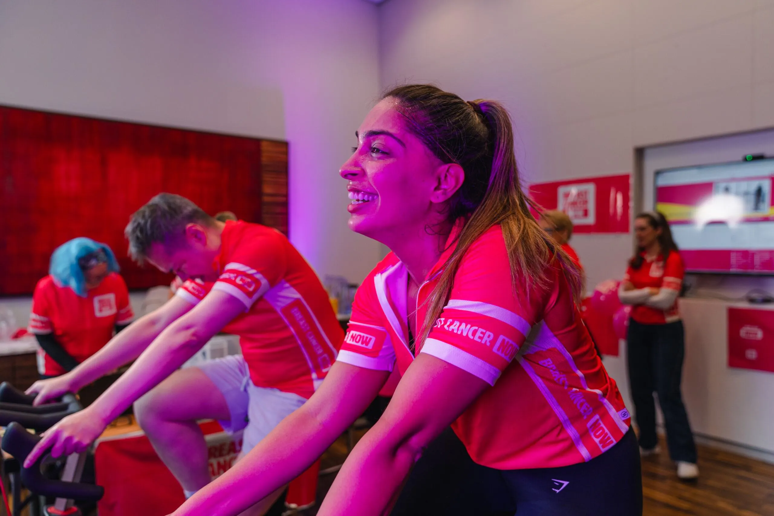 A group of people in pink cycling jerseys participate in a stationary cycling session indoors. In the foreground, a smiling woman with a ponytail pedals a bike under pink-purple lighting, while others ride behind her and a few stand watching in the background.