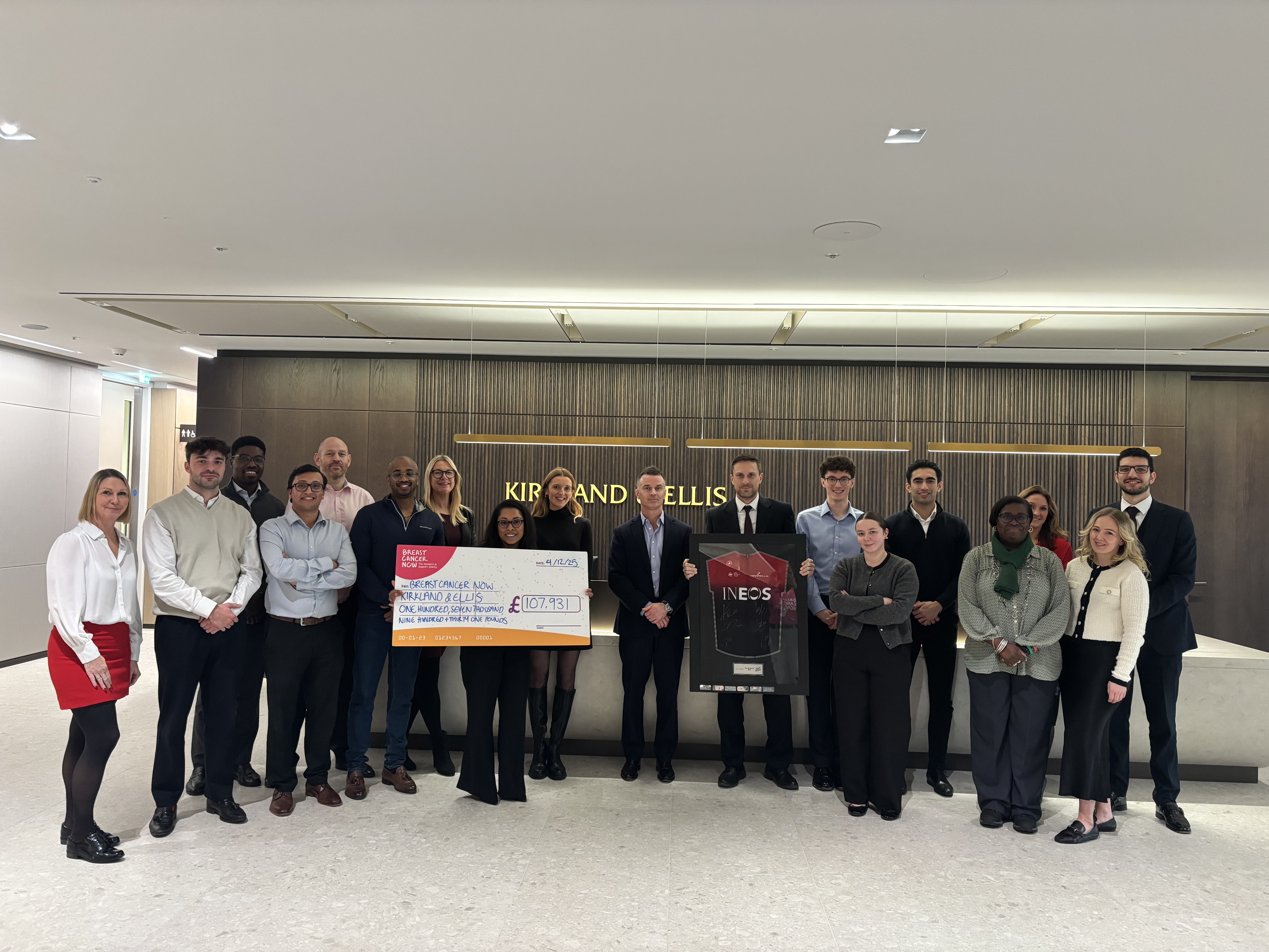 A group of colleagues are standing together, formally dressed holding the Tour de Law prize, a framed Team INEOS Grenadiers jersey and a giant cheque detailing they’ve raised £107,931 for Breast Cancer Now. 