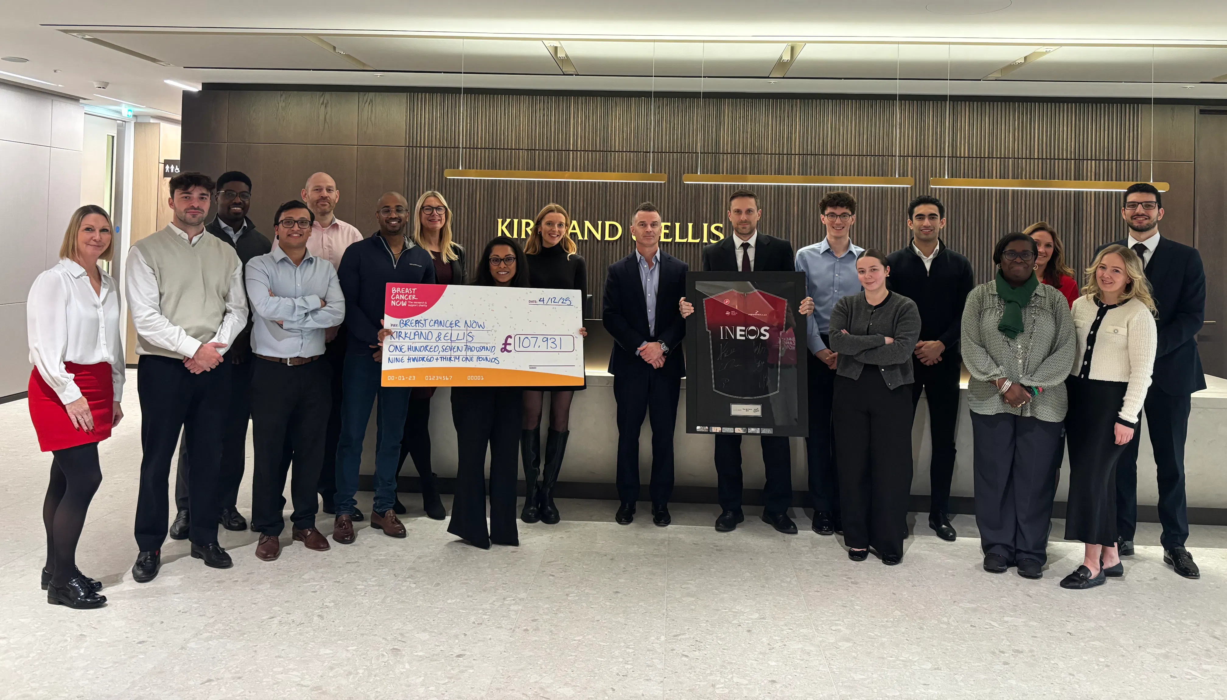 A group of colleagues are standing together, formally dressed holding the Tour de Law prize, a framed Team INEOS Grenadiers jersey and a giant cheque detailing they’ve raised £107,931 for Breast Cancer Now.