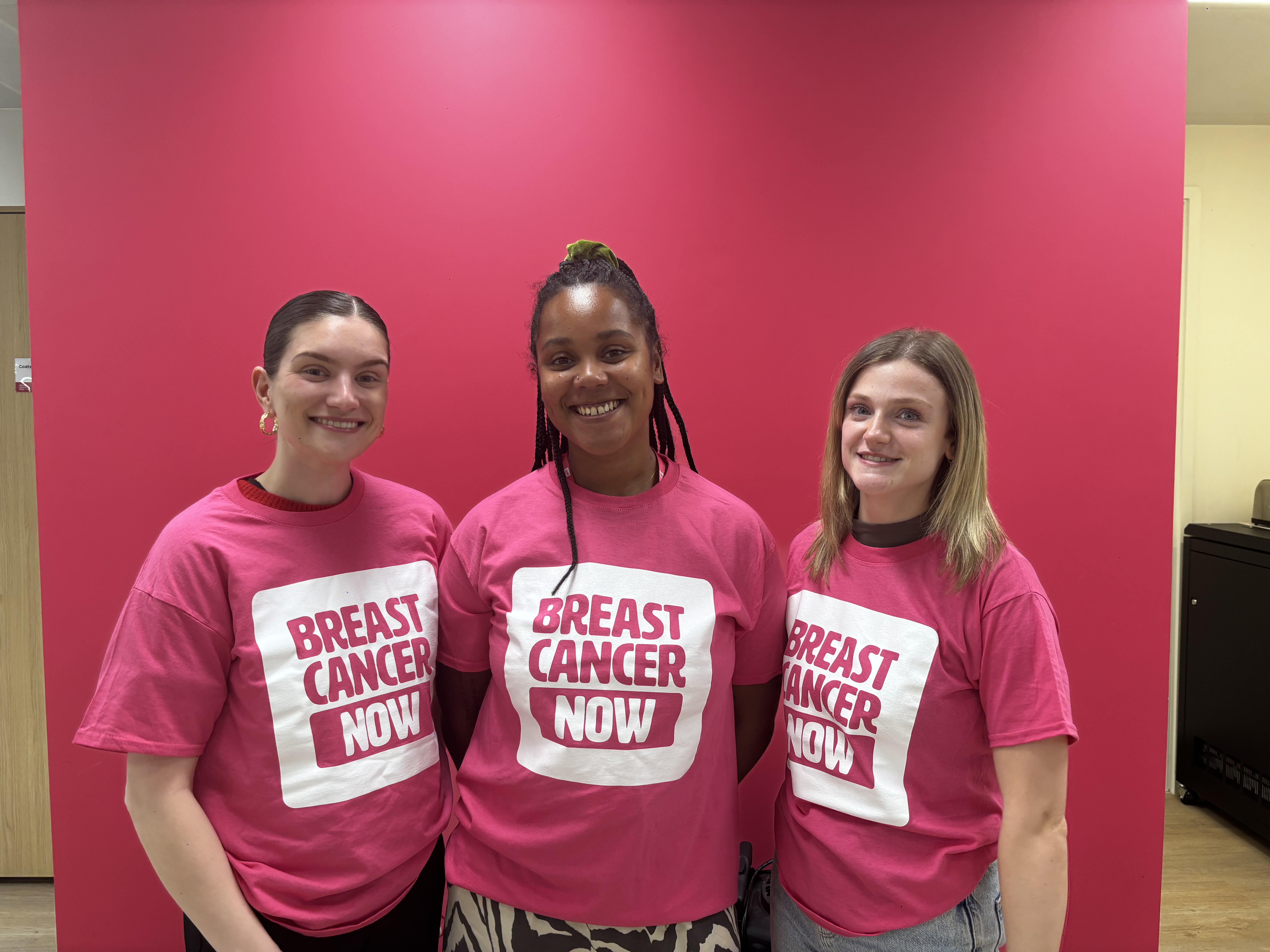 Three women are standing close together, smiling, wearing pink “Breast Cancer Now” t-shirts in front of a pink background. 