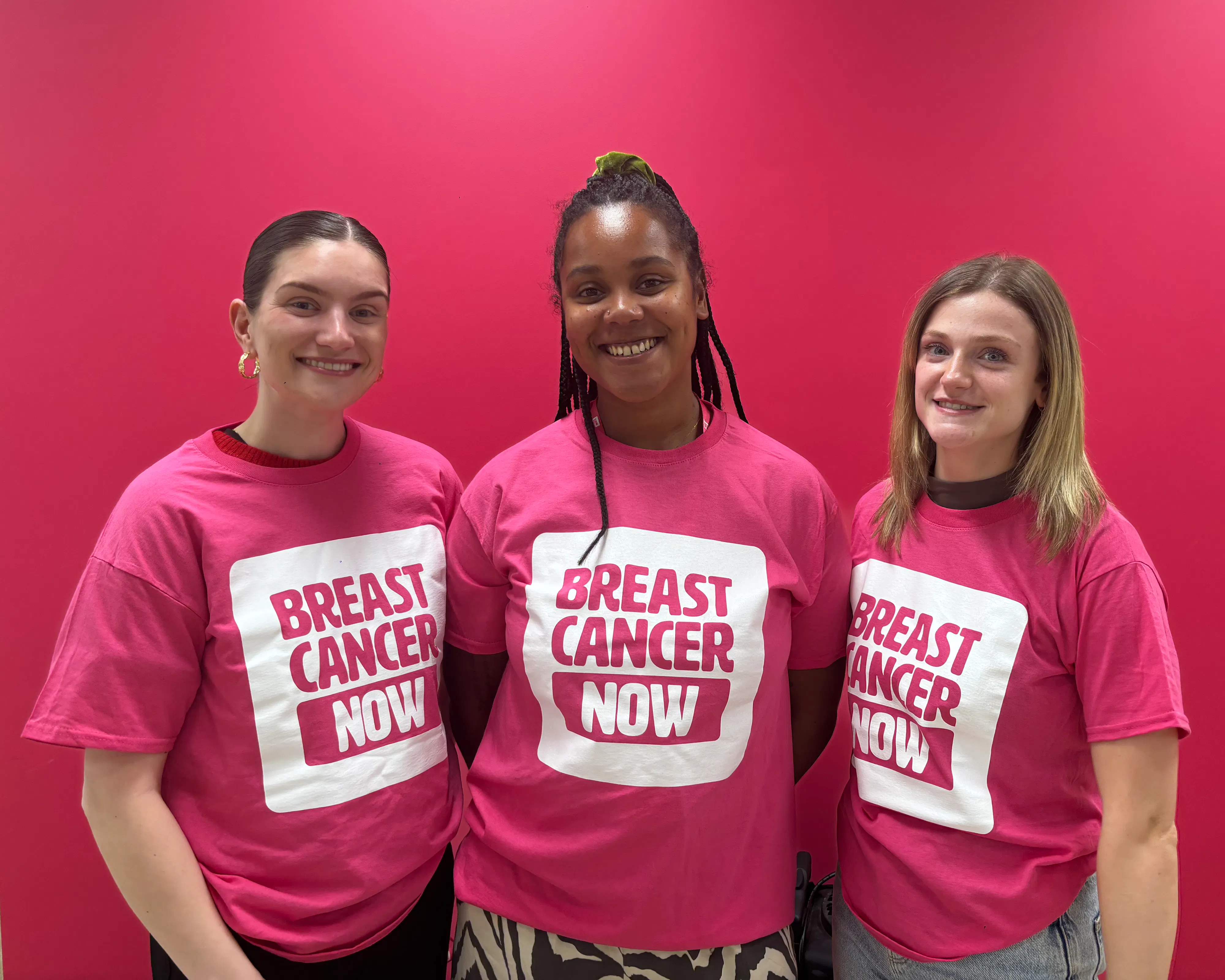 Three women are standing close together, smiling, wearing pink “Breast Cancer Now” t-shirts in front of a pink background.