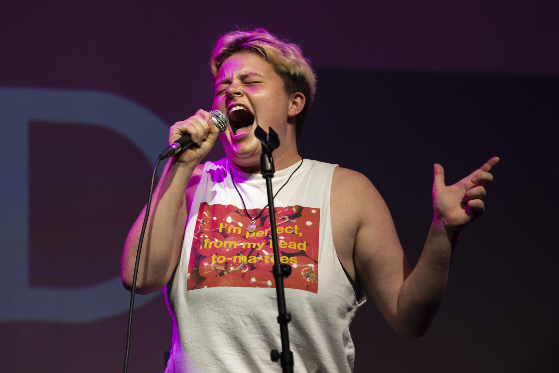 A participant performing on stage dressed in a vest and shouting into the microphone with their eyes shut.