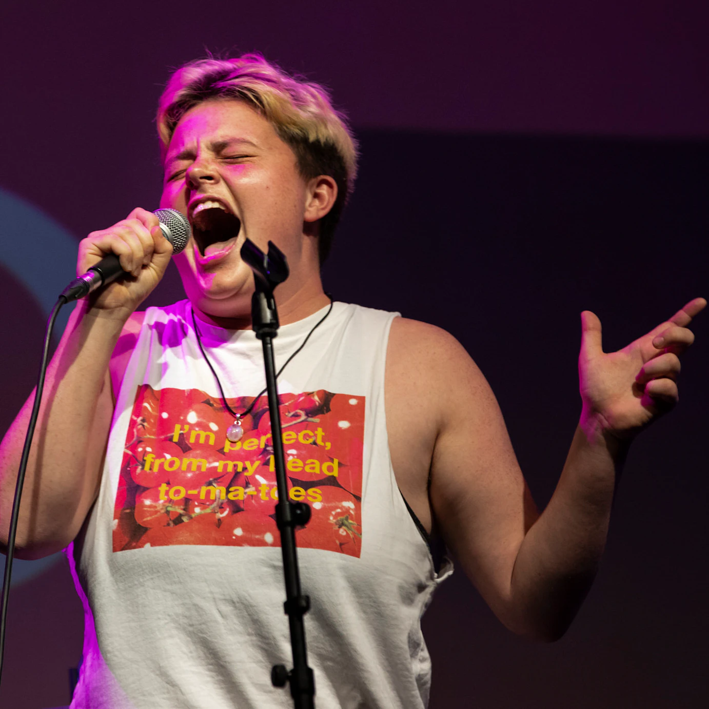 A participant performing on stage dressed in a vest and shouting into the microphone with their eyes shut.