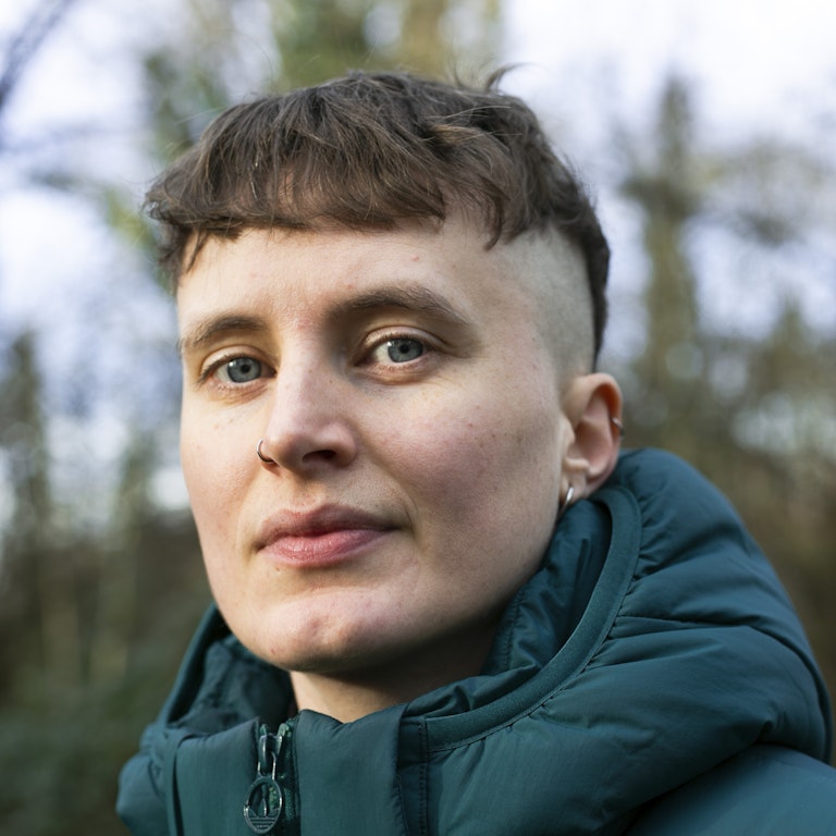 A headshot of the artist I. JORDAN. They are wearing a dark green coat with trees behind.