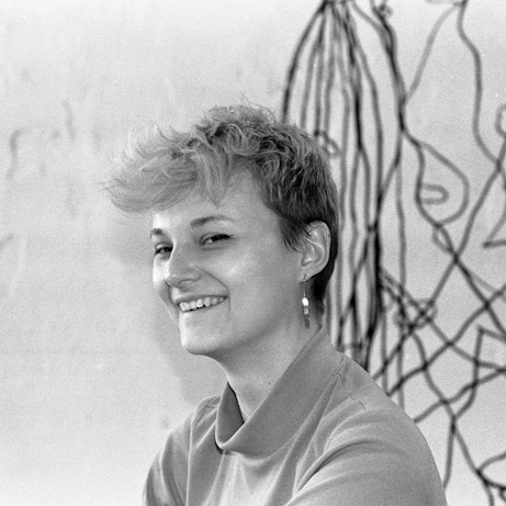 Sonia Killmann smiling into the camera. Behind them falling from above are what look like tangled black wires. The photo is tinted black and white.