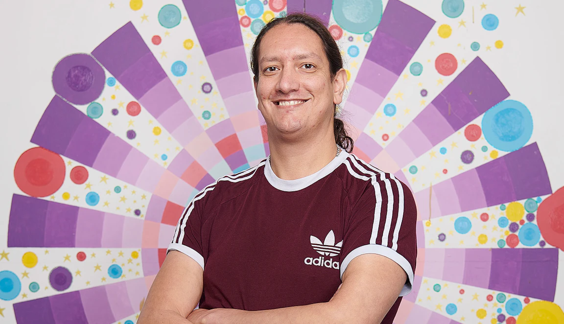 Leon Patel stood with his arms crossed smiling into the camera. Behind him is a multicoloured circular pattern painted on a white wall.