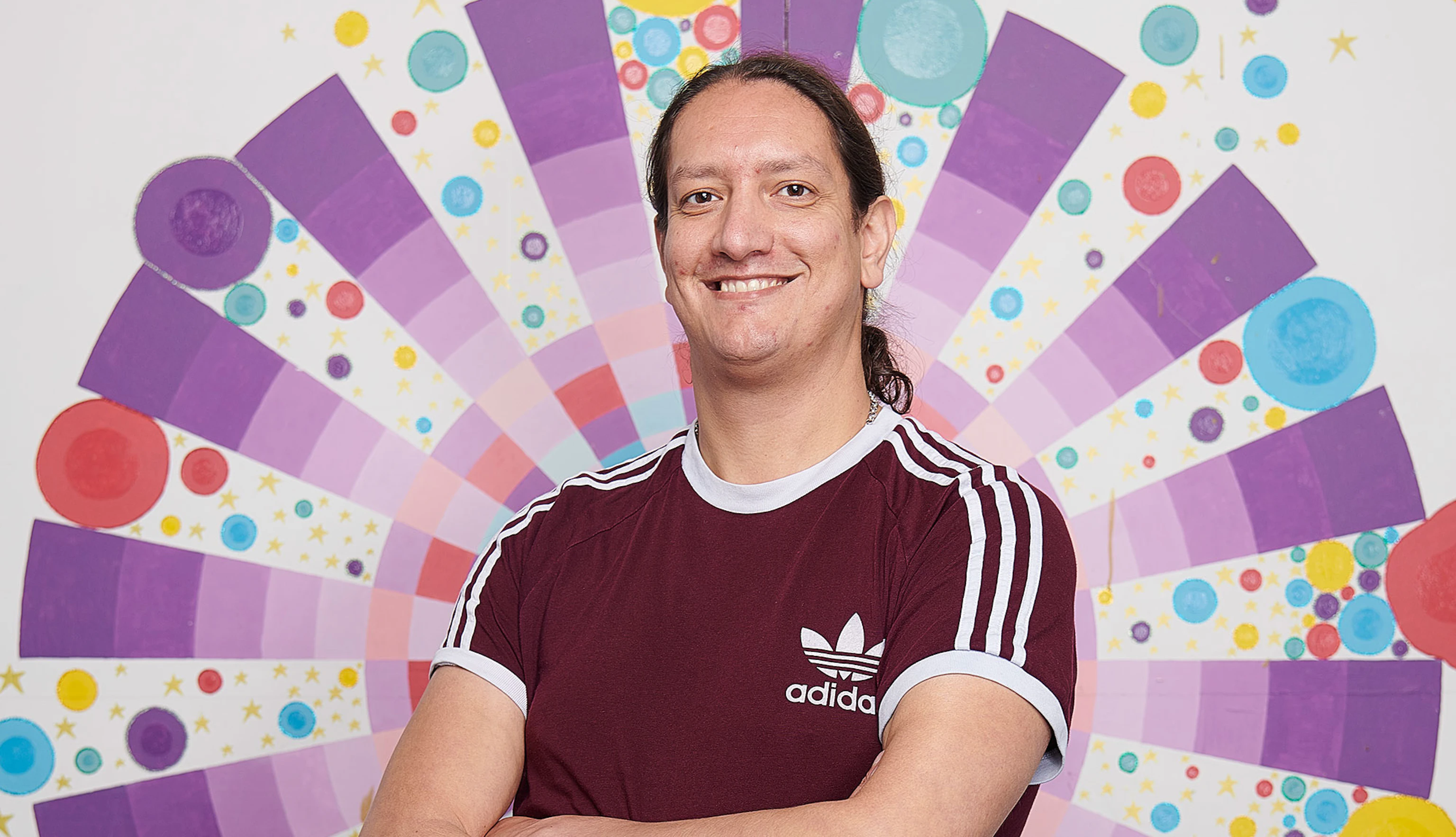 Leon Patel stood with his arms crossed smiling into the camera. Behind him is a multicoloured circular pattern painted on a white wall.