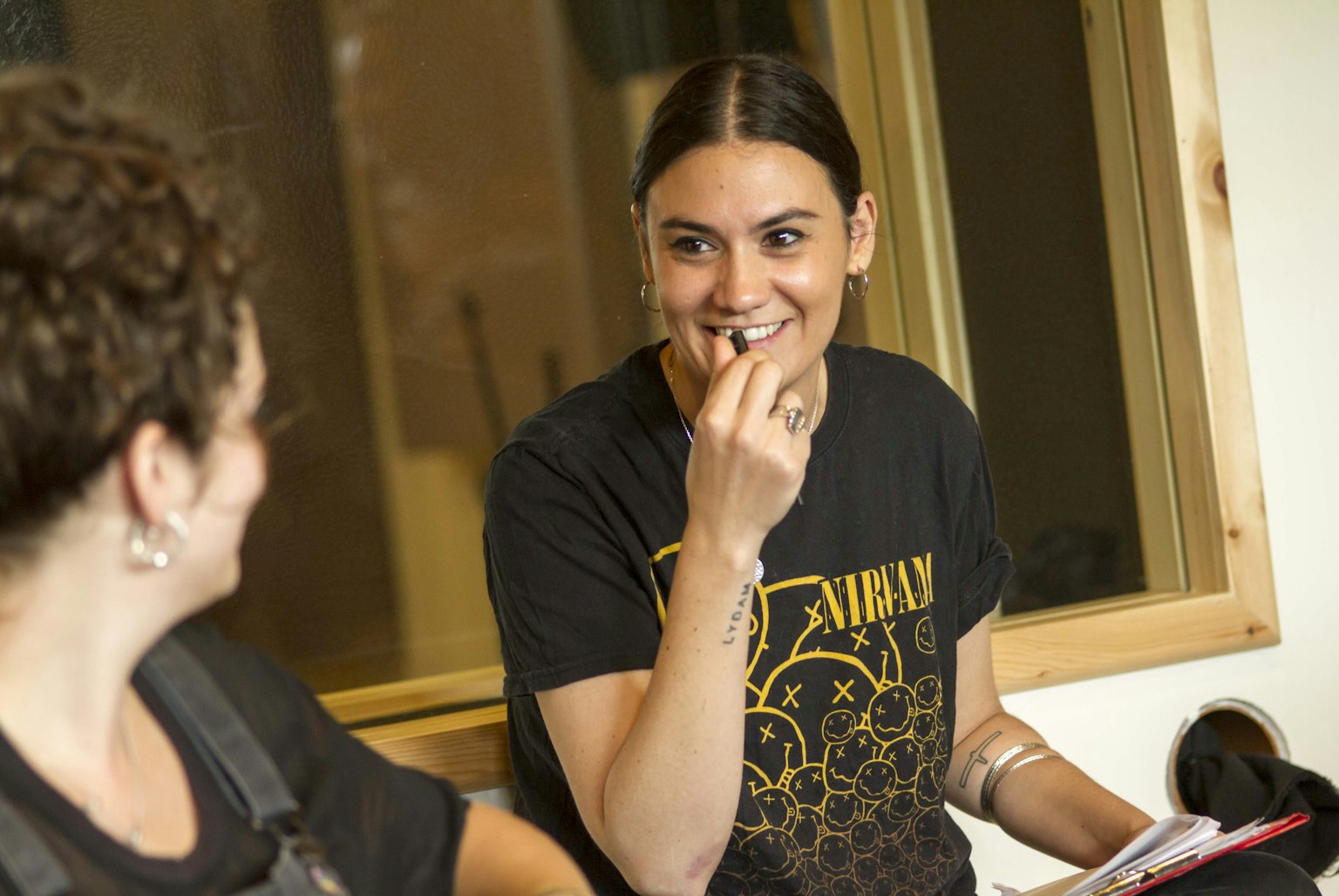 The artist Nadine Shah is smiling at a participant. Nadine is wearing a black Nirvana t-shirt. They are both sitting down in front of a window. 