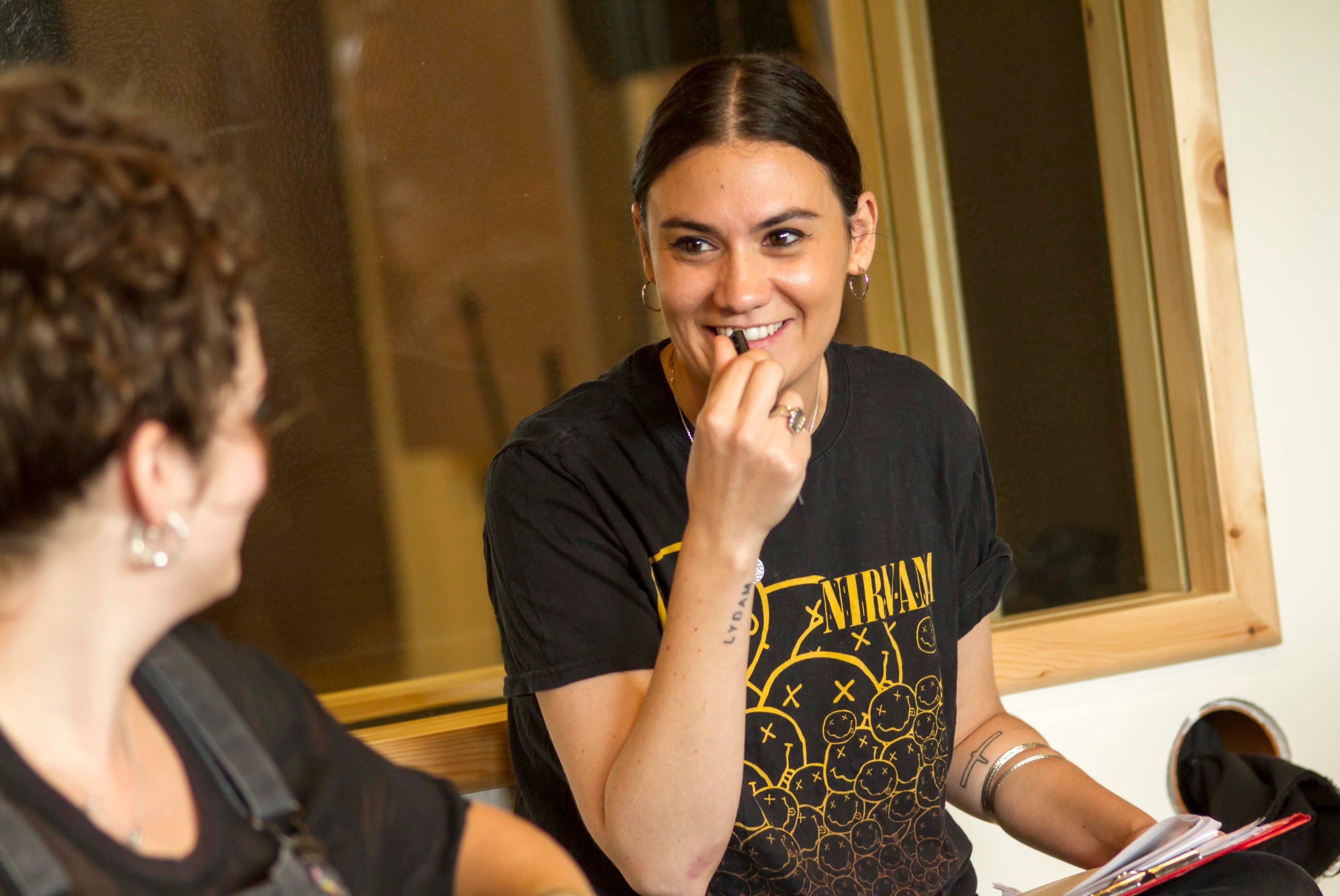 The artist Nadine Shah is smiling at a participant. Nadine is wearing a black Nirvana t-shirt. They are both sitting down in front of a window.