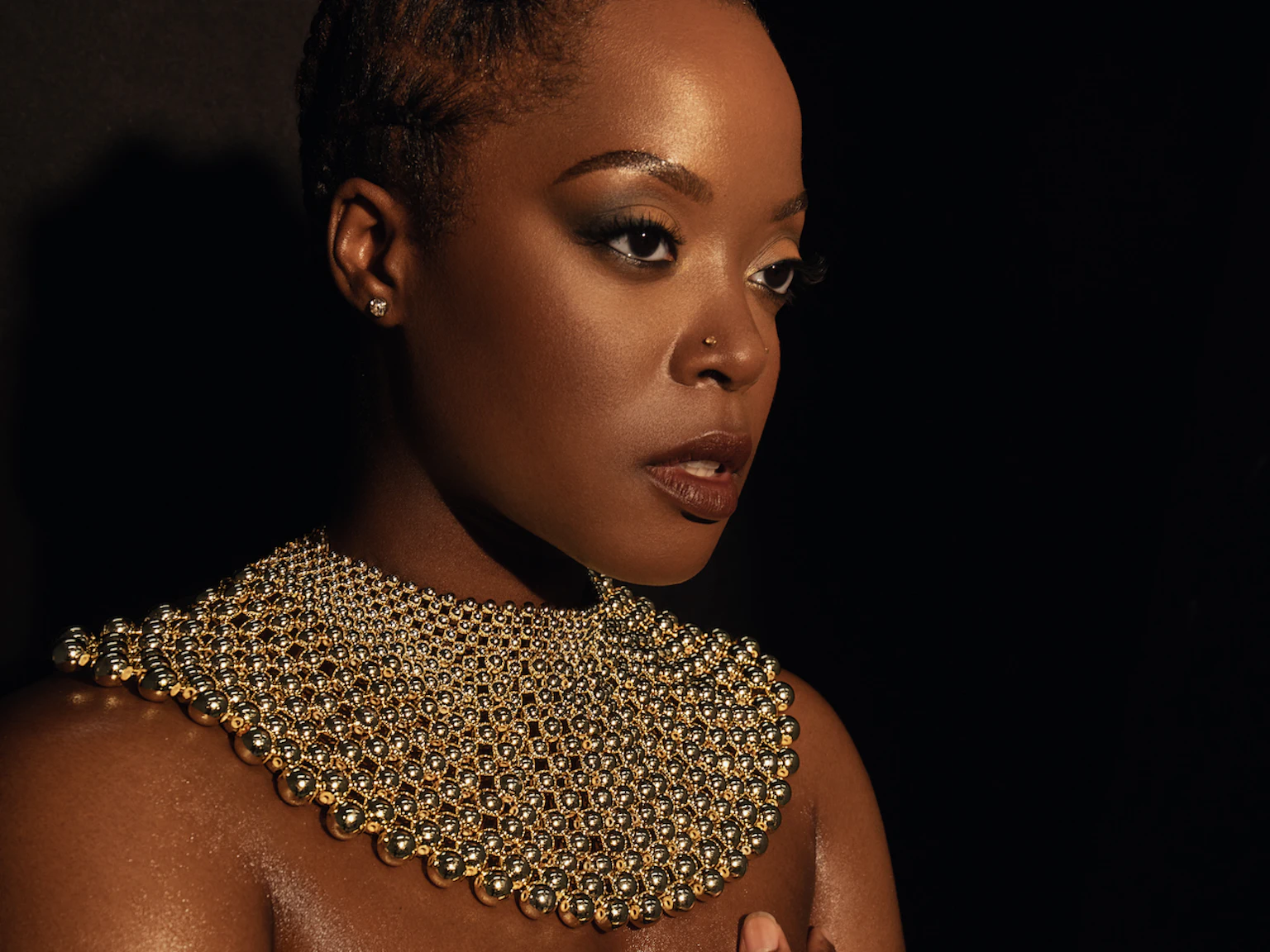 Karen Nyame KG dressed in black with a loose gold choker necklace made from beads, and bracelet. One hand is over her chest as she stares off pensively into the distance.