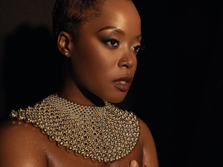Karen Nyame KG dressed in black with a loose gold choker necklace made from beads, and bracelet. One hand is over her chest as she stares off pensively into the distance.