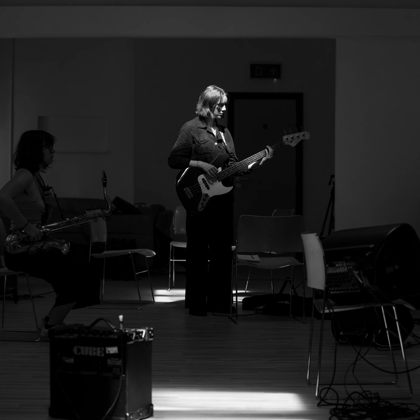 The rehearsal room. Most of the room is in darkness except a bassist in the centre lit by the sunlight seeping in through ceiling window.