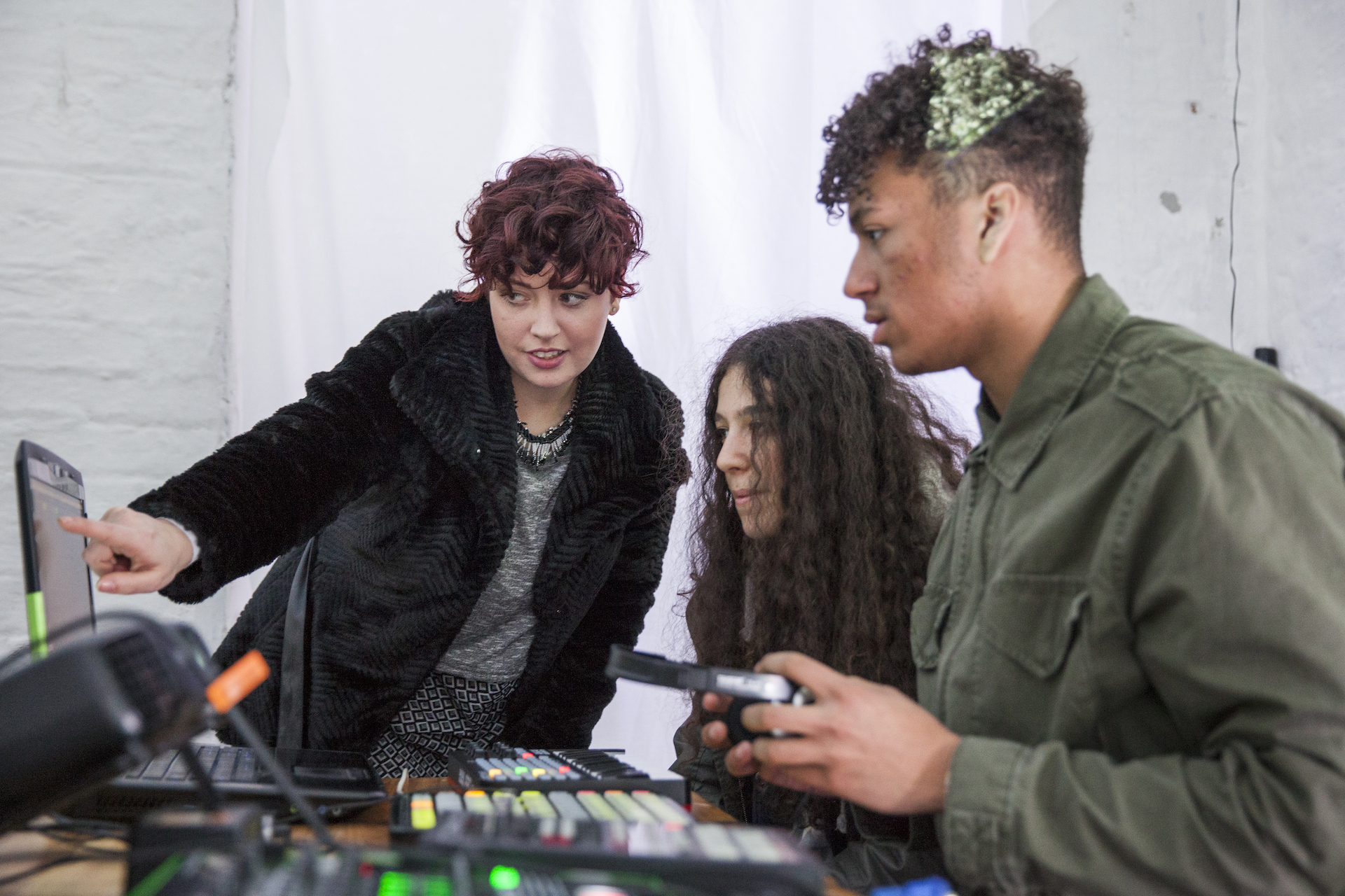 Two participants look at a laptop screen, the table is filled with production equipment. The facilitator is pointing to the screen. 