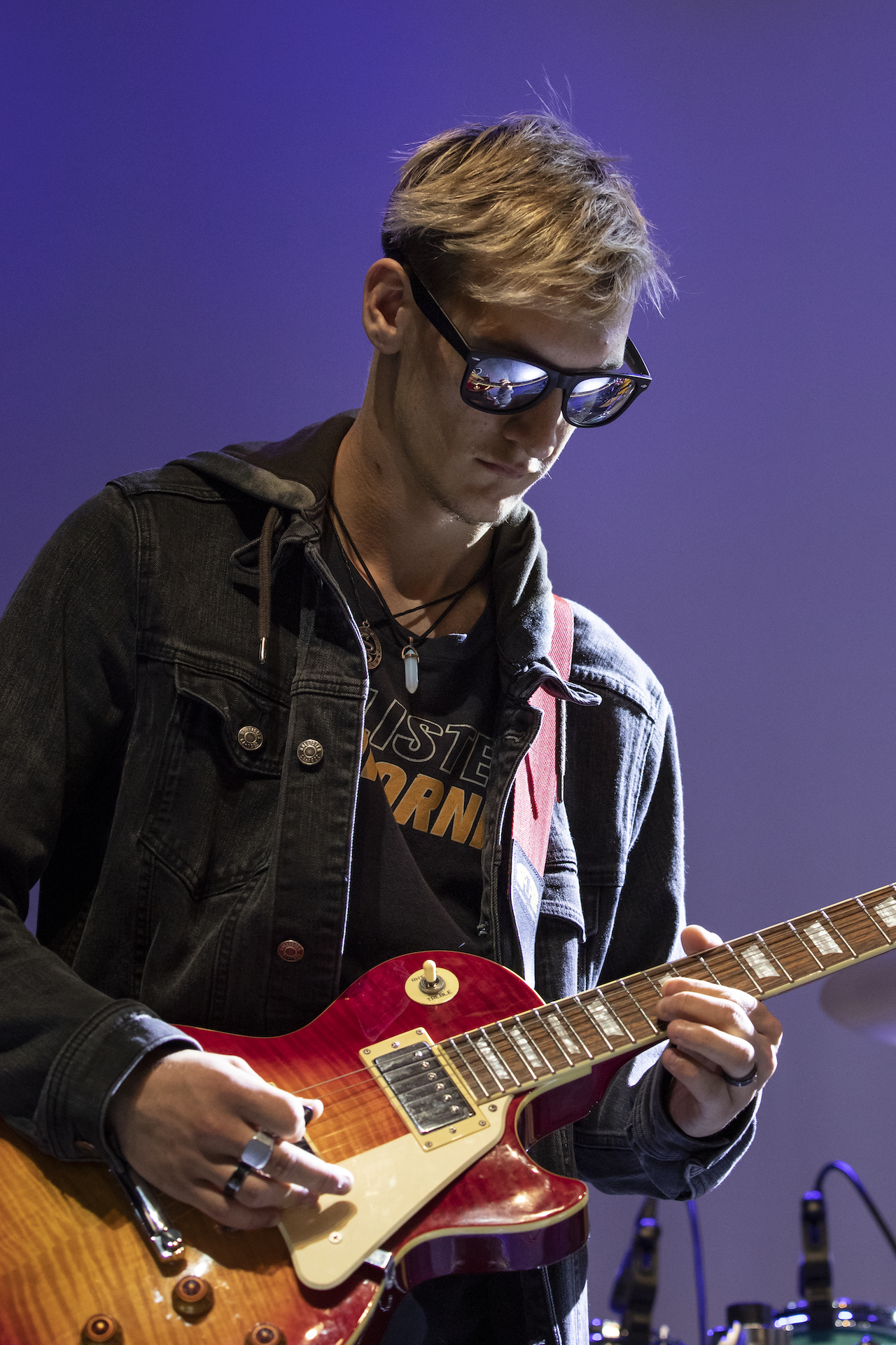 A participant playing guitar on stage wearing a denim jacket and sunglasses that reflect the stage.