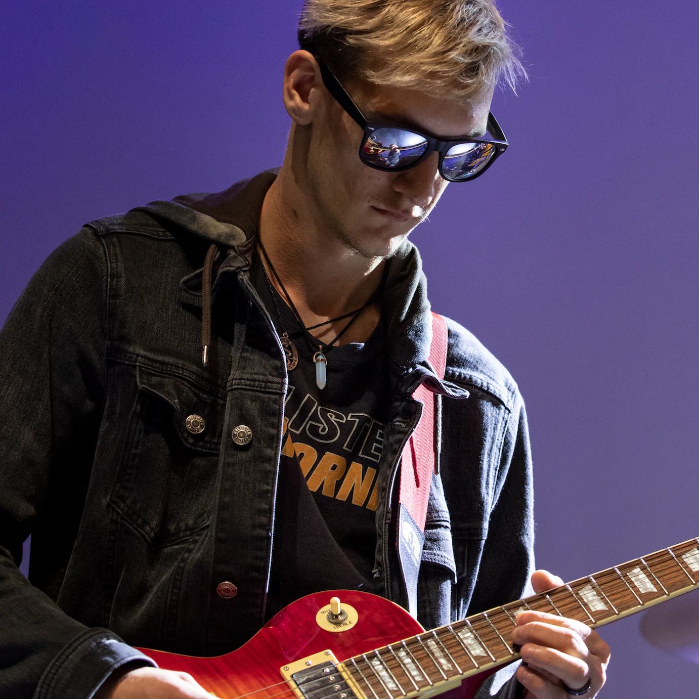 A participant playing guitar on stage wearing a denim jacket and sunglasses that reflect the stage.