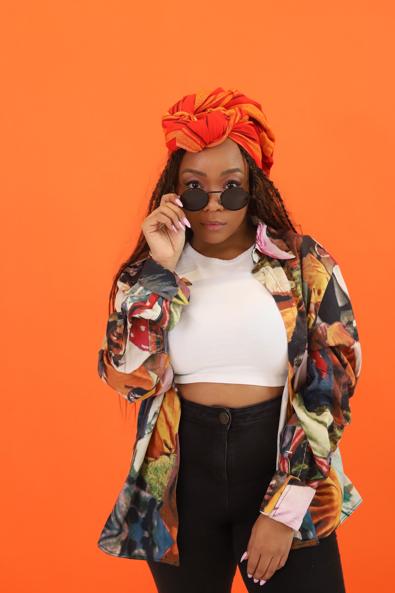 Artist Lebo standing against a bright orange backdrop. They are wearing a brightly coloured shirt and orange headscarf, looking directly at the camera with shades lowered.
