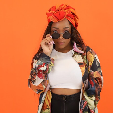 Artist Lebo standing against a bright orange backdrop. They are wearing a brightly coloured shirt and orange headscarf, looking directly at the camera with shades lowered.