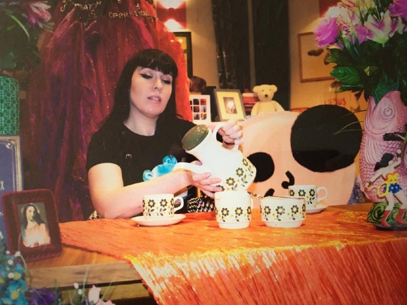 Clare MuNulty sat by a table with a shiny orange throw on the top. On it sits four white teacups with flower patterns along the side. Clare is holding a teapot with the same patterns. Behind them are various other bits and pieces like flowers, picture frames and a teddy bear.