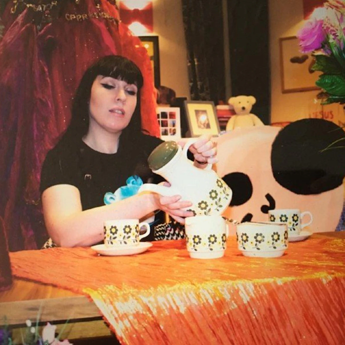 Clare MuNulty sat by a table with a shiny orange throw on the top. On it sits four white teacups with flower patterns along the side. Clare is holding a teapot with the same patterns. Behind them are various other bits and pieces like flowers, picture frames and a teddy bear.