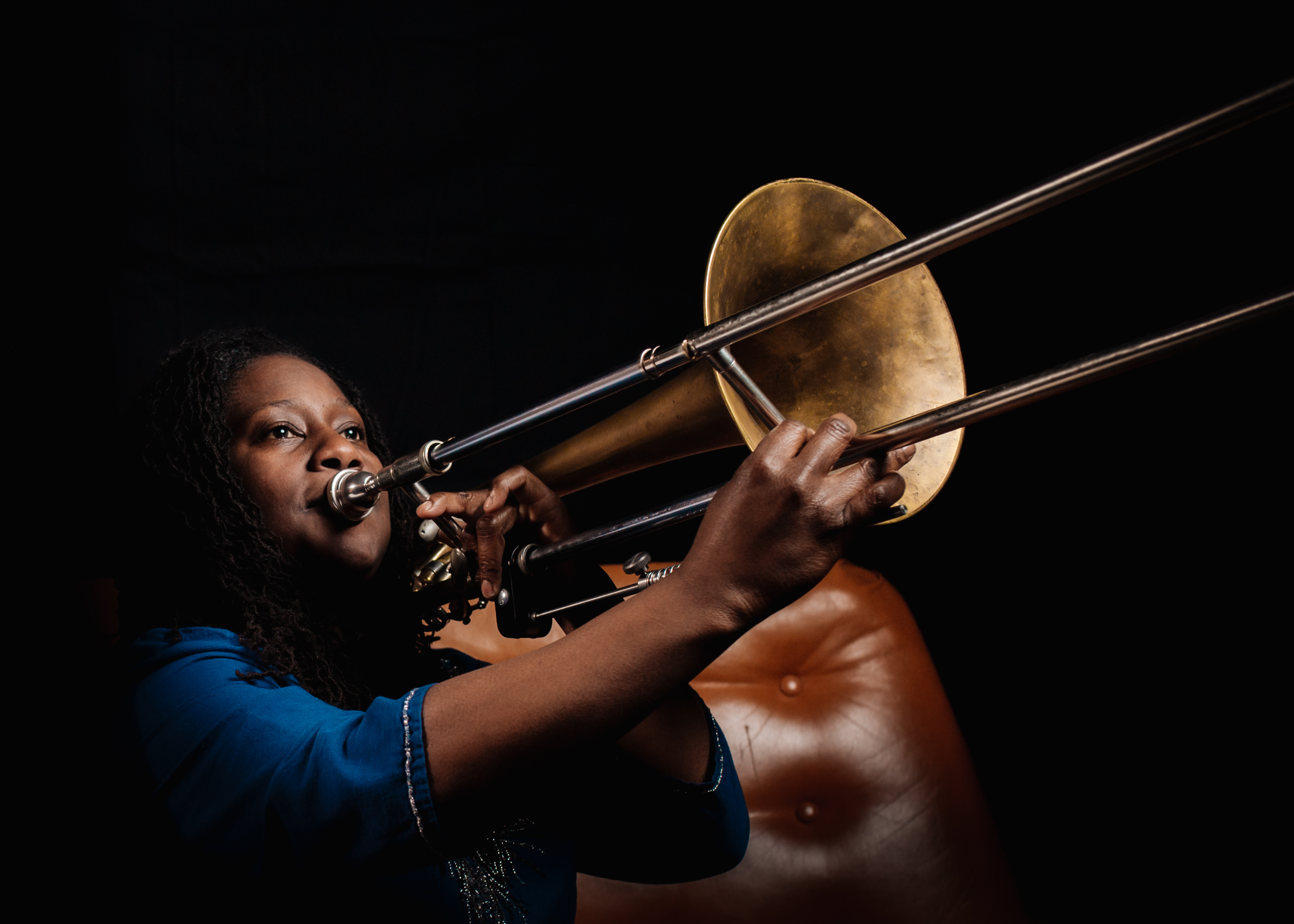 Hannabiell Sanders playing the trombone in a dark space with a light brown leather couch beside her.