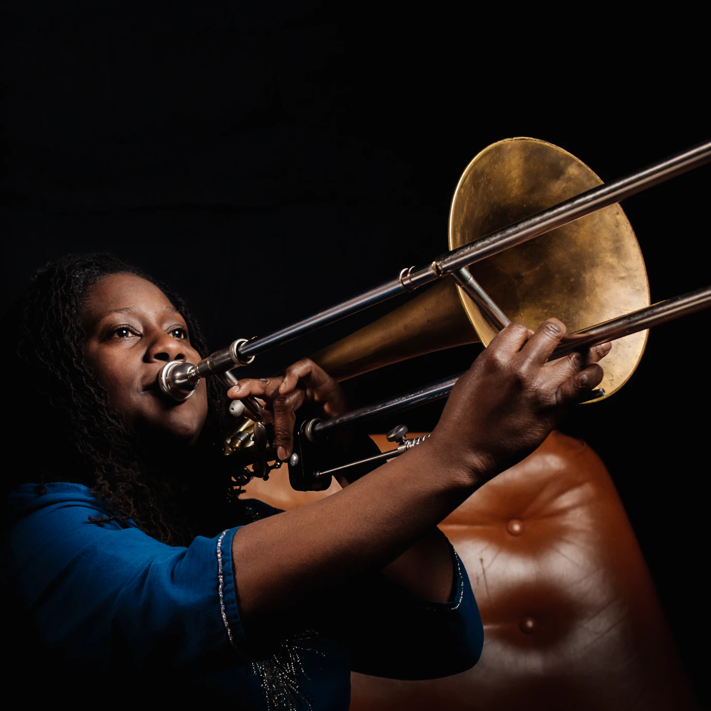 Hannabiell Sanders playing the trombone in a dark space with a light brown leather couch beside her.