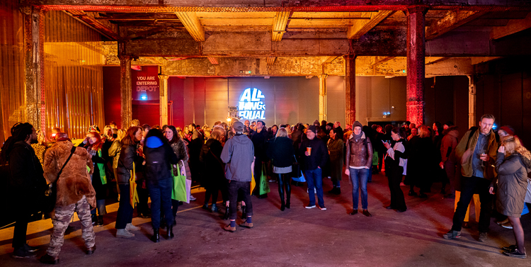 Crowds of people gathering within the Depot Mayfield for our Manifesto Launch. The dark room is illuminated by red and yellow lighting cast on the walls and pillars.