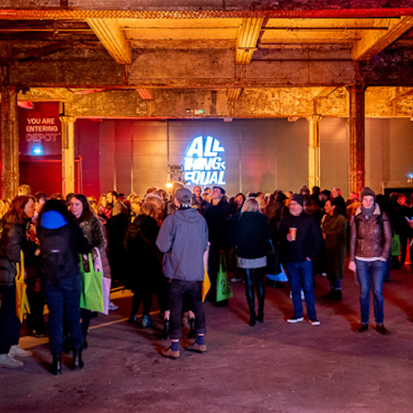 Crowds of people gathering within the Depot Mayfield for our Manifesto Launch. The dark room is illuminated by red and yellow lighting cast on the walls and pillars.